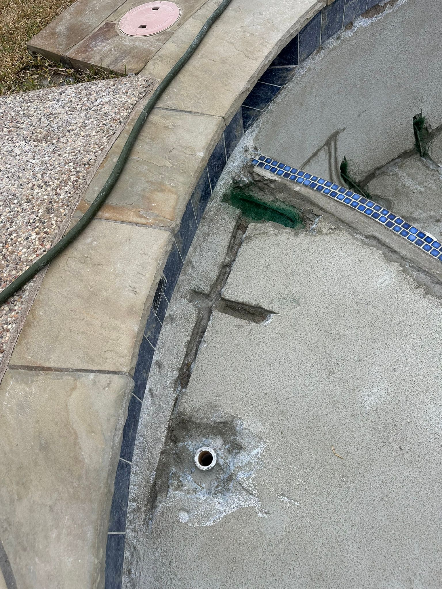 Overhead view of a concrete pool edge with a drain, mosaic tiles, and a green hose.
