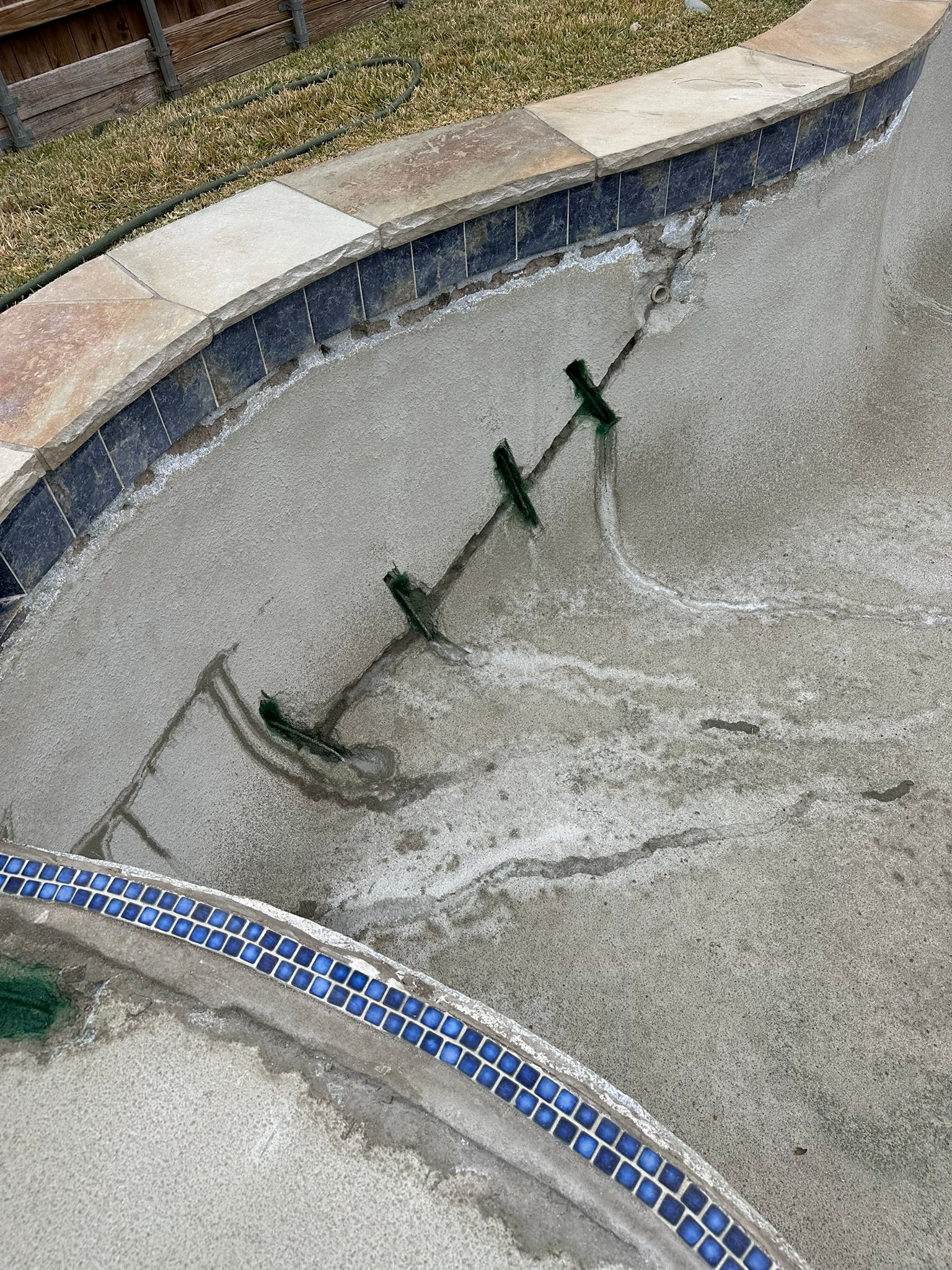 Empty pool with visible cracks and green fixtures in the concrete. Blue tile border on the edge.