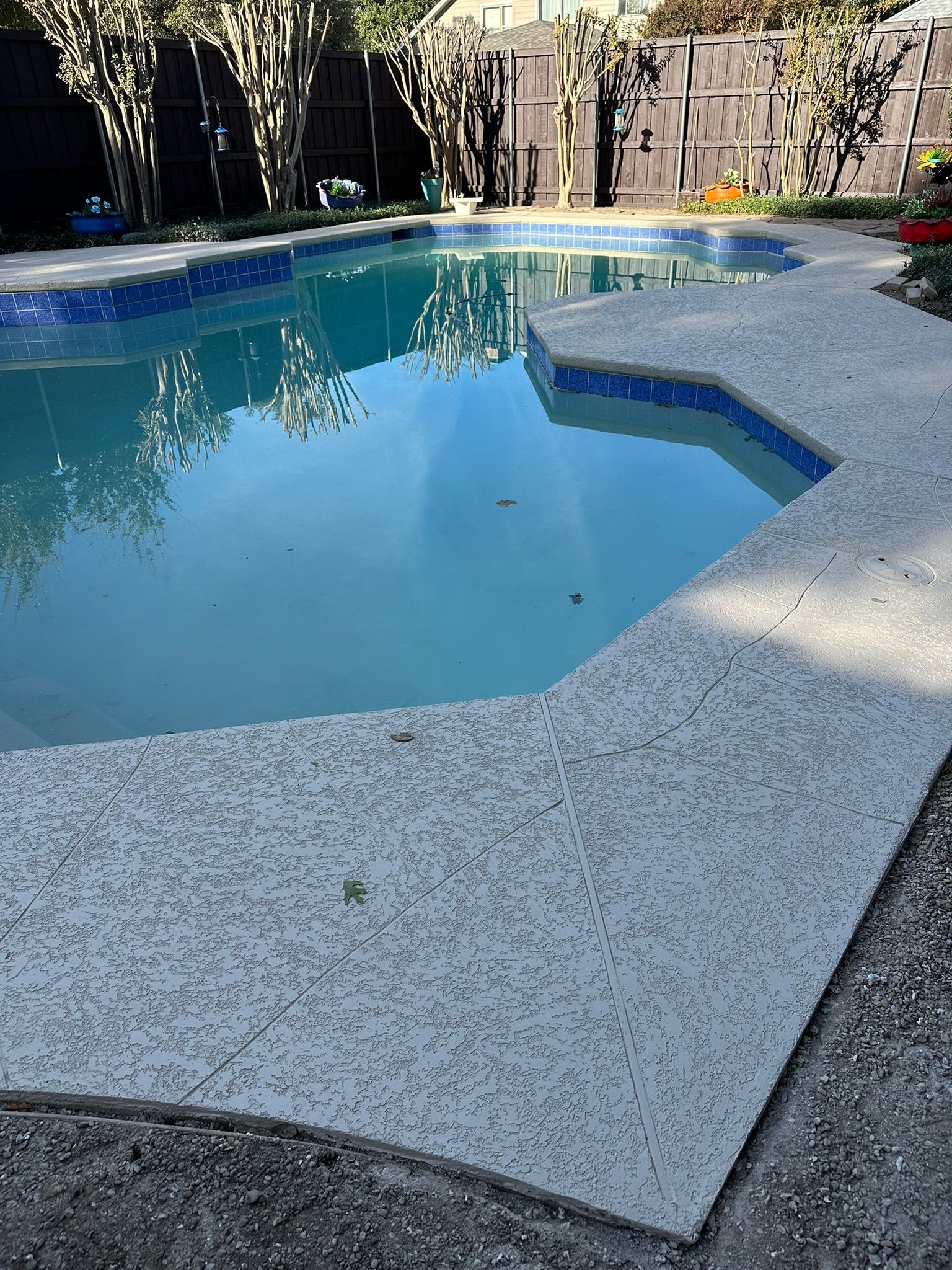 Swimming pool with gray speckled concrete decking. Blue water reflects sky. Fenced backyard.