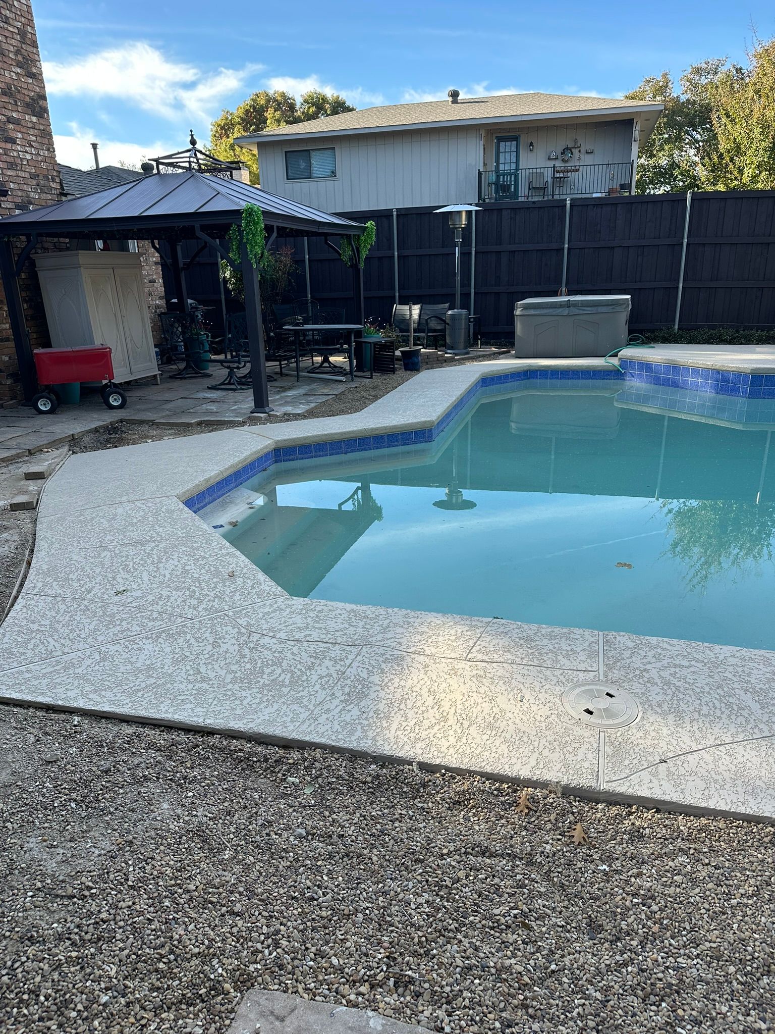 Pool with concrete border, blue tiles, patio furniture, hot tub, and dark fence in background.