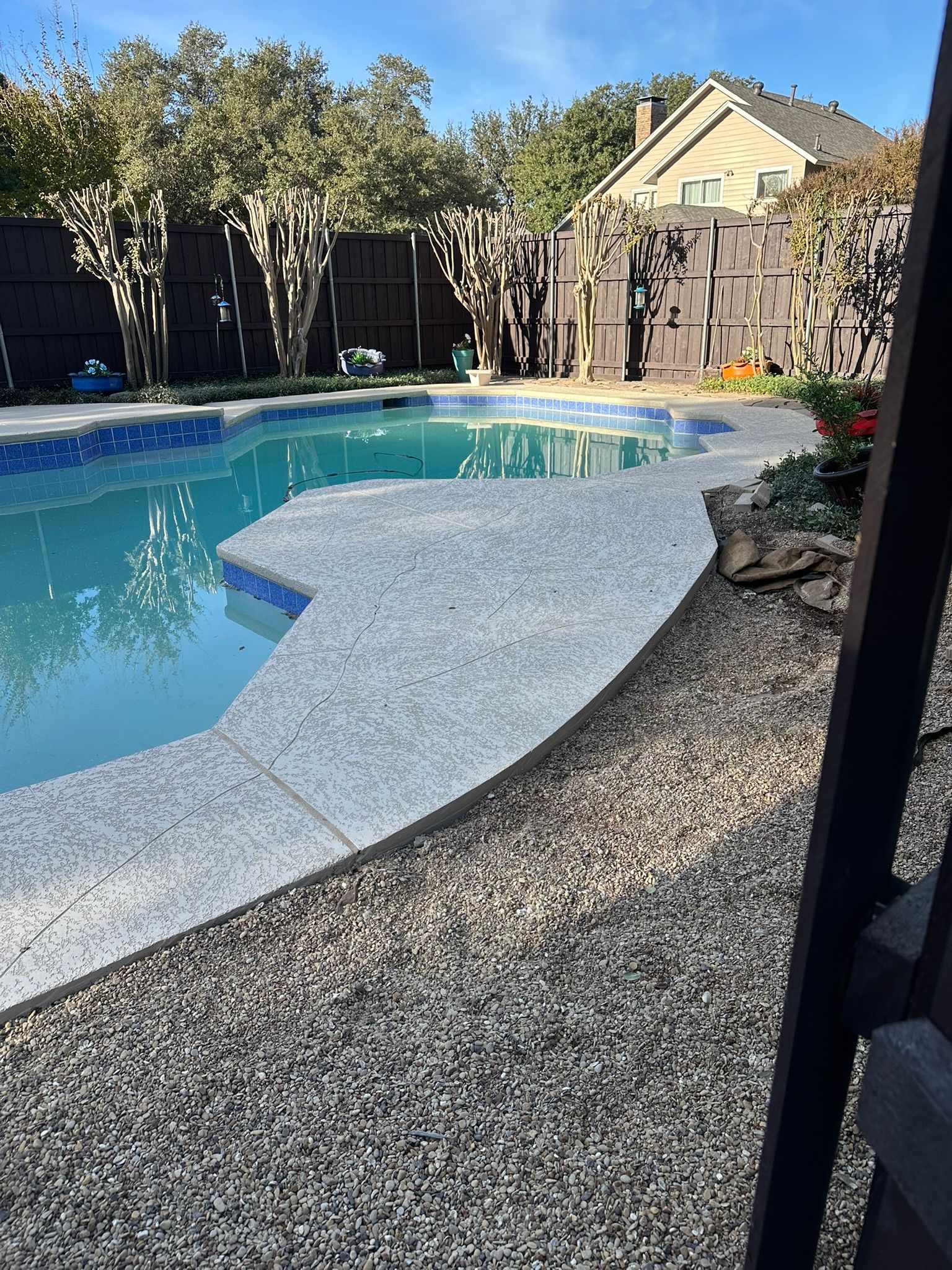 Pool area with concrete patio, blue water, dark fence, and trees.