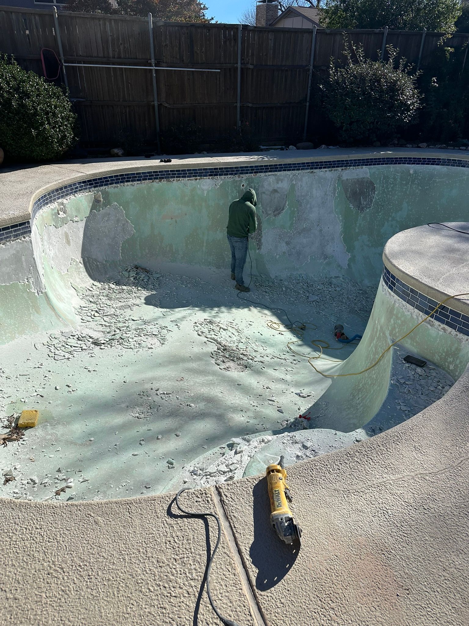 Person in green suit working in an empty, algae-covered swimming pool with tool. Concrete patio and wooden fence in background.