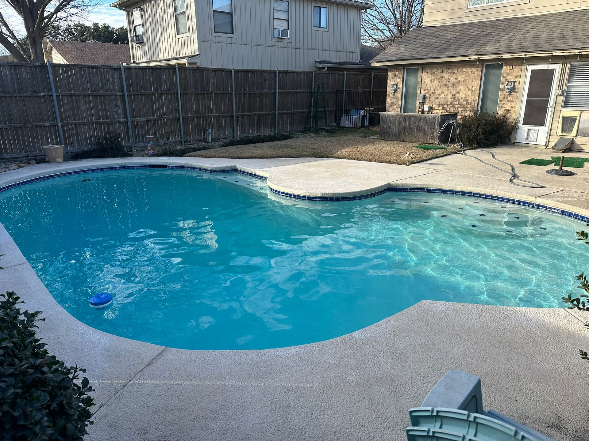 Pool with turquoise water surrounded by a concrete patio. A house and wooden fence are in the background.