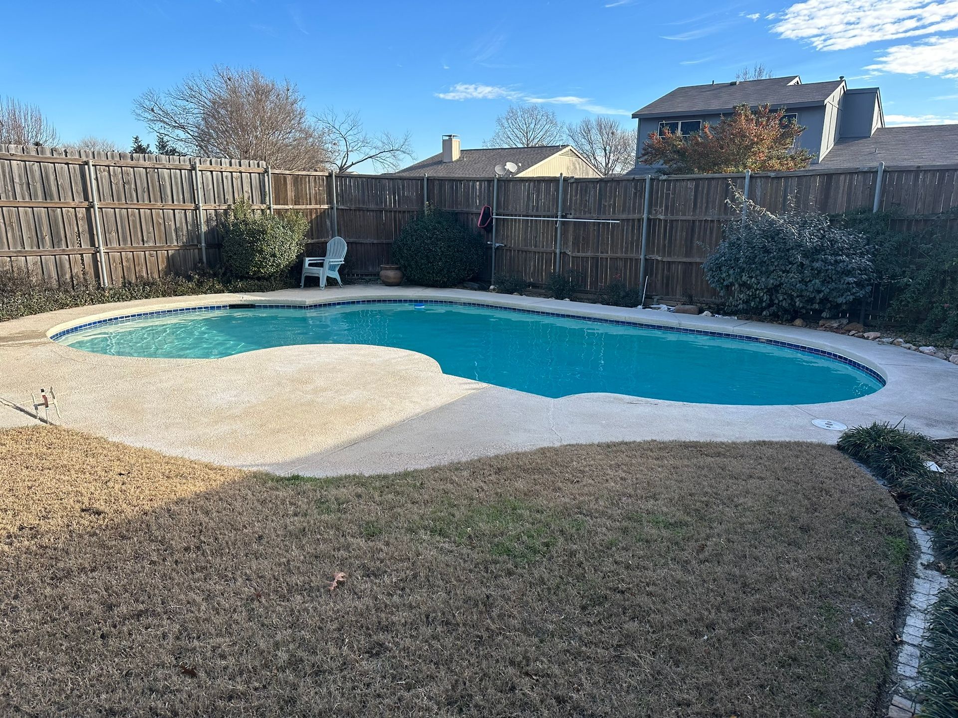 Backyard pool with tan concrete deck, brown fence, and dead grass. Clear blue sky.