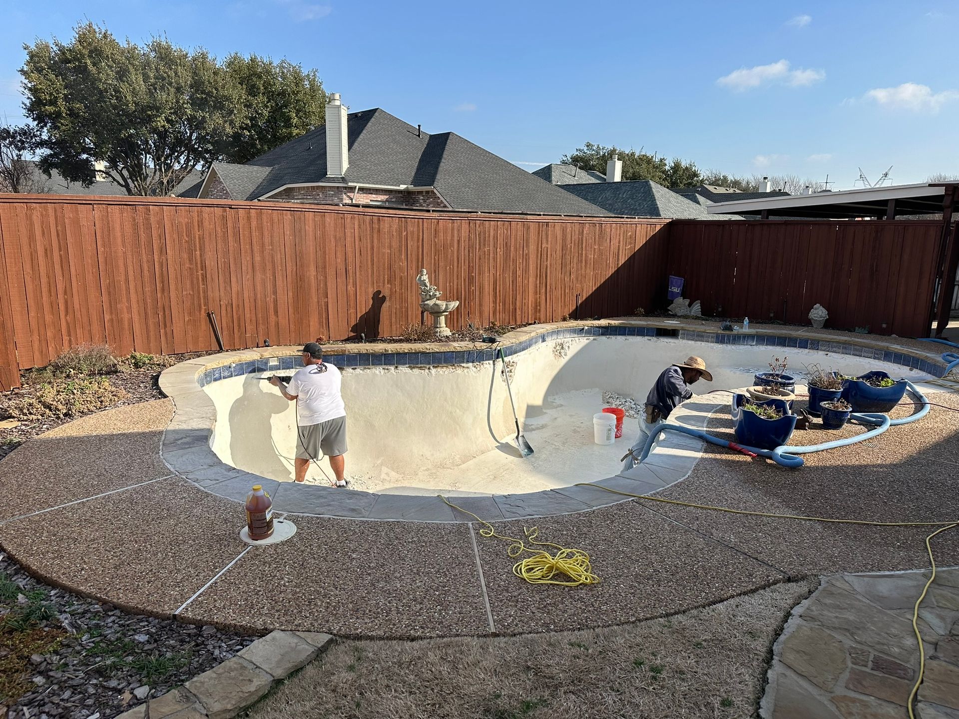 Two people working on a drained pool. They are in a backyard with a brown fence on a sunny day.