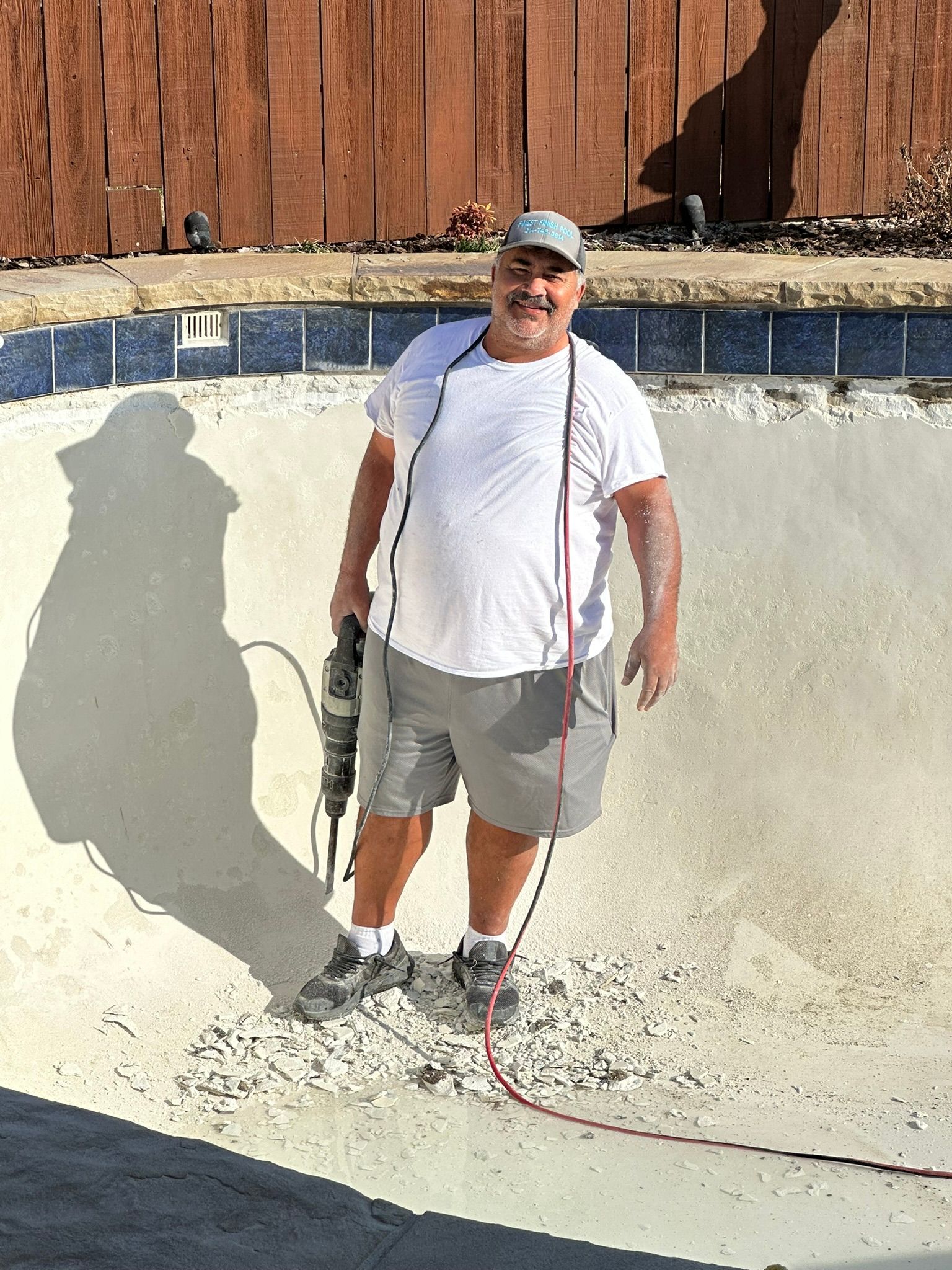 Man standing in an empty pool, holding a drill. He wears a white shirt, gray shorts, and a cap.