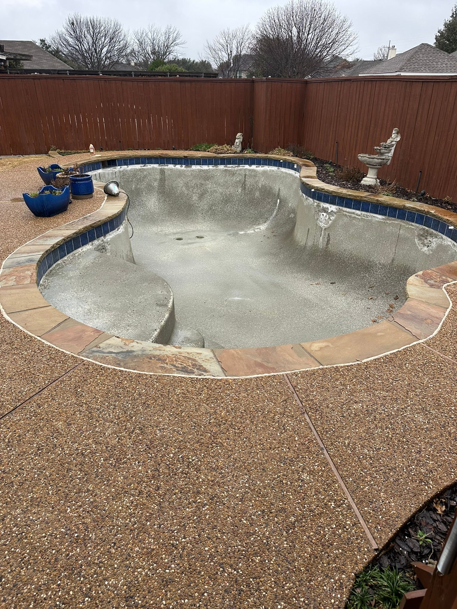 Empty, kidney-shaped pool surrounded by brown brick and stone, set in a backyard with a brick wall.