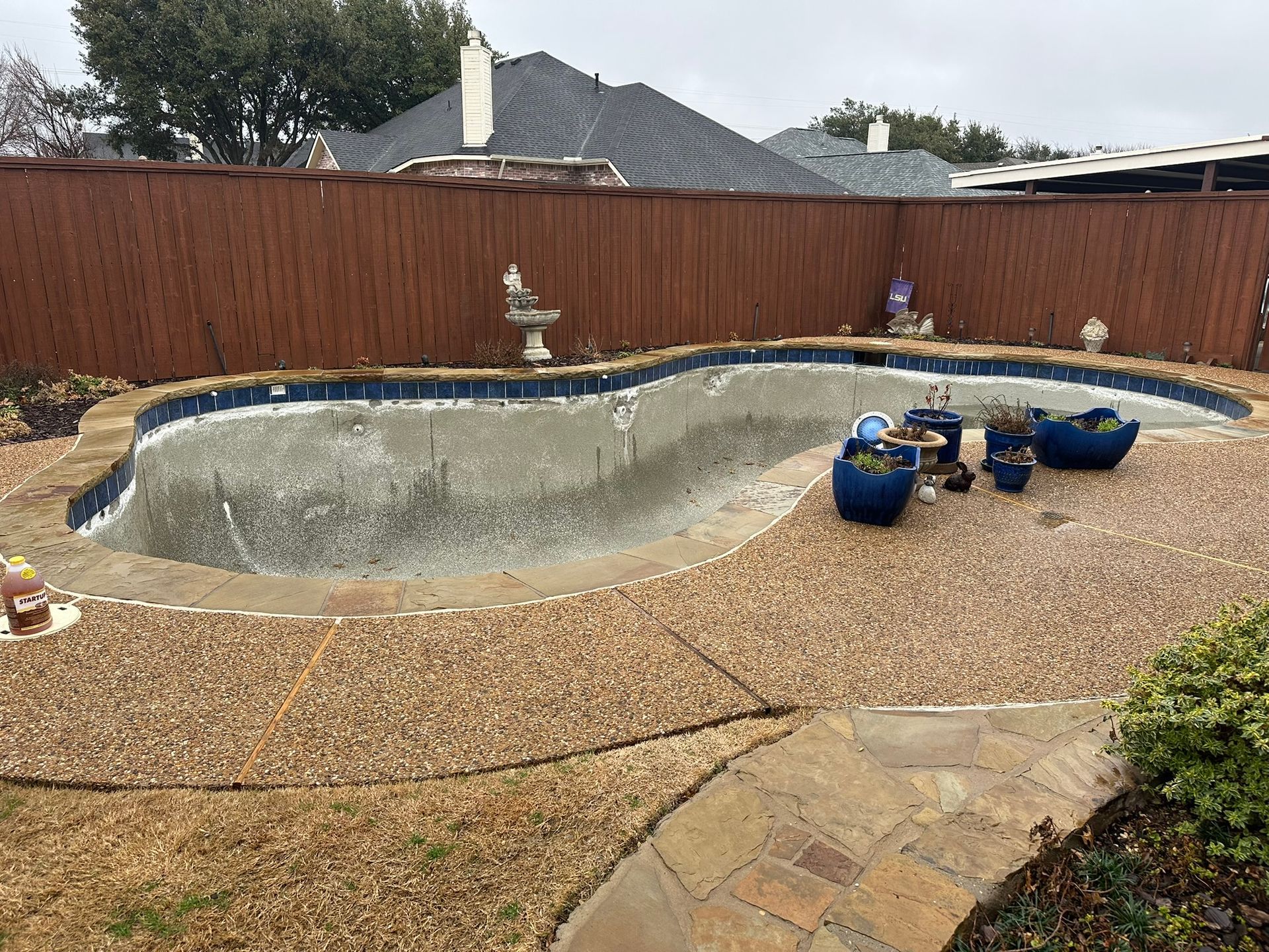 Empty, oval-shaped swimming pool in a backyard, surrounded by gravel and a brown wooden fence.