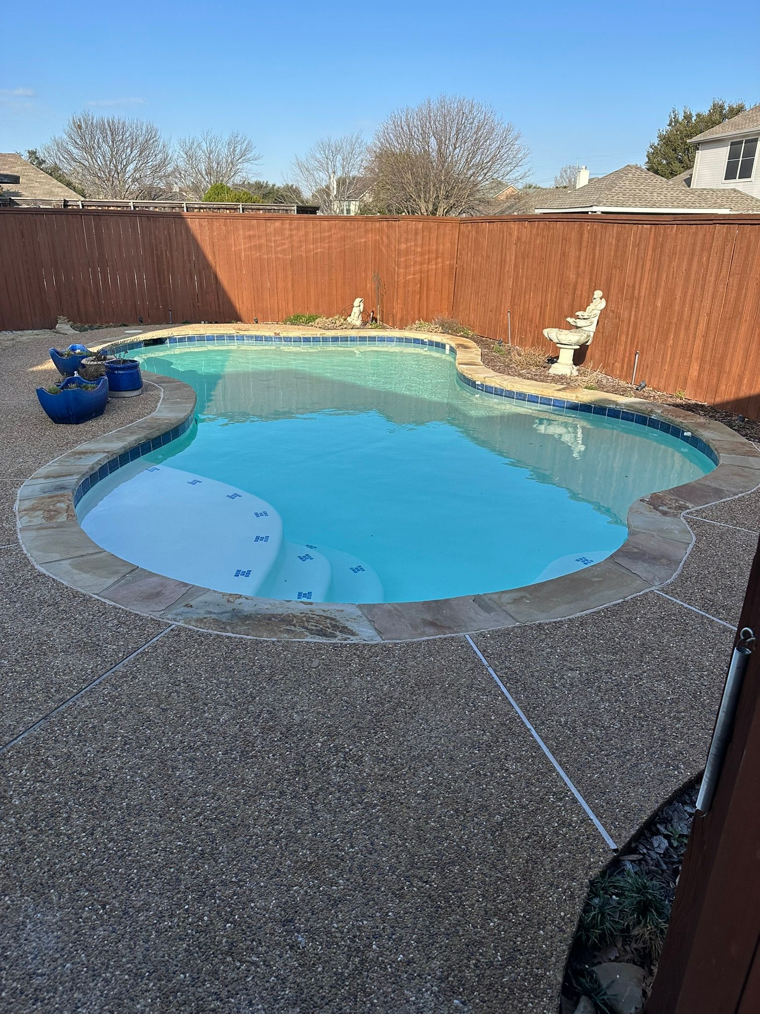 A kidney-shaped swimming pool surrounded by concrete, with a wood fence in the background, on a sunny day.