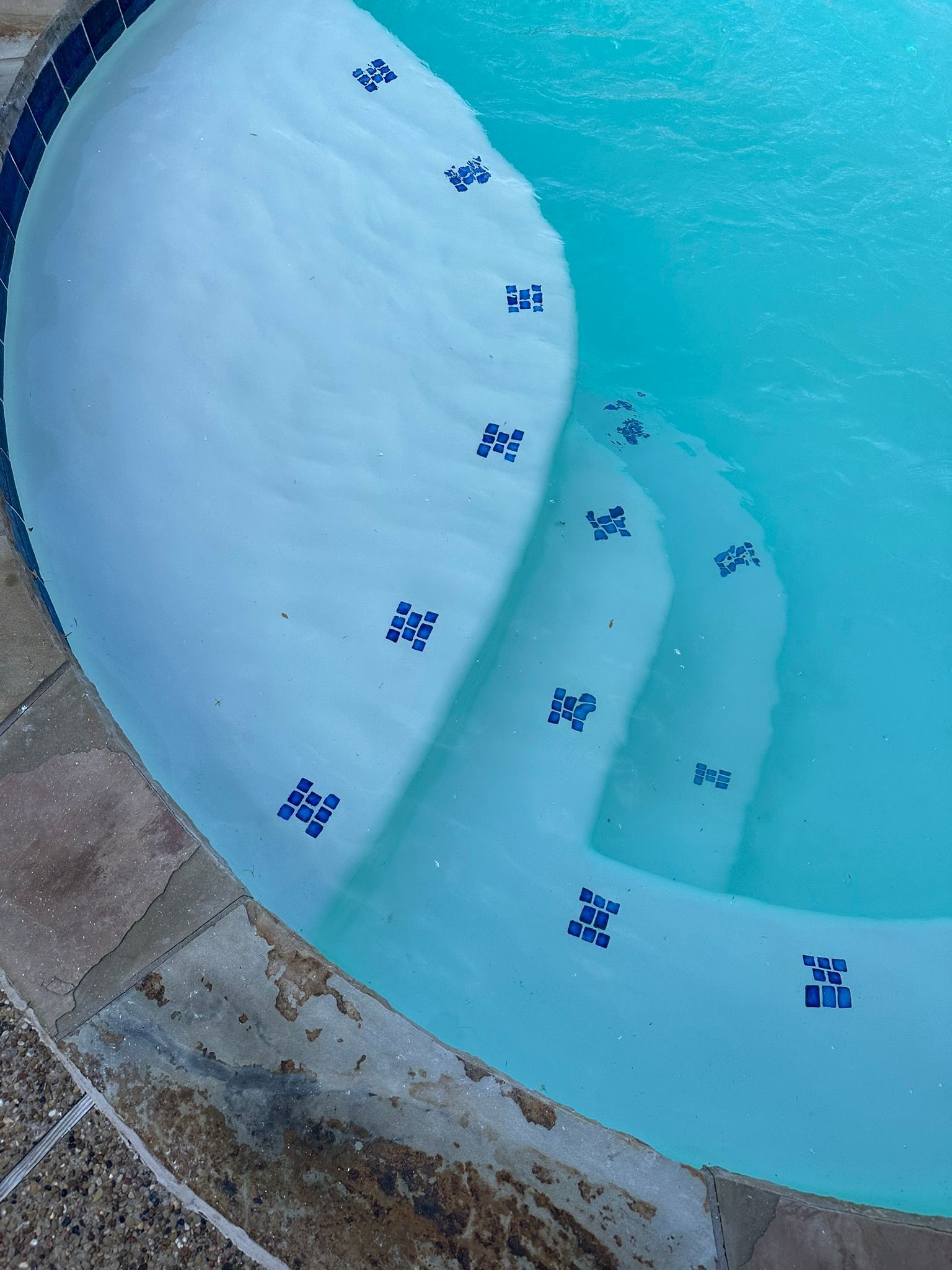 Pool steps with blue tile detail, submerged in clear, turquoise water. Stone coping surrounds.