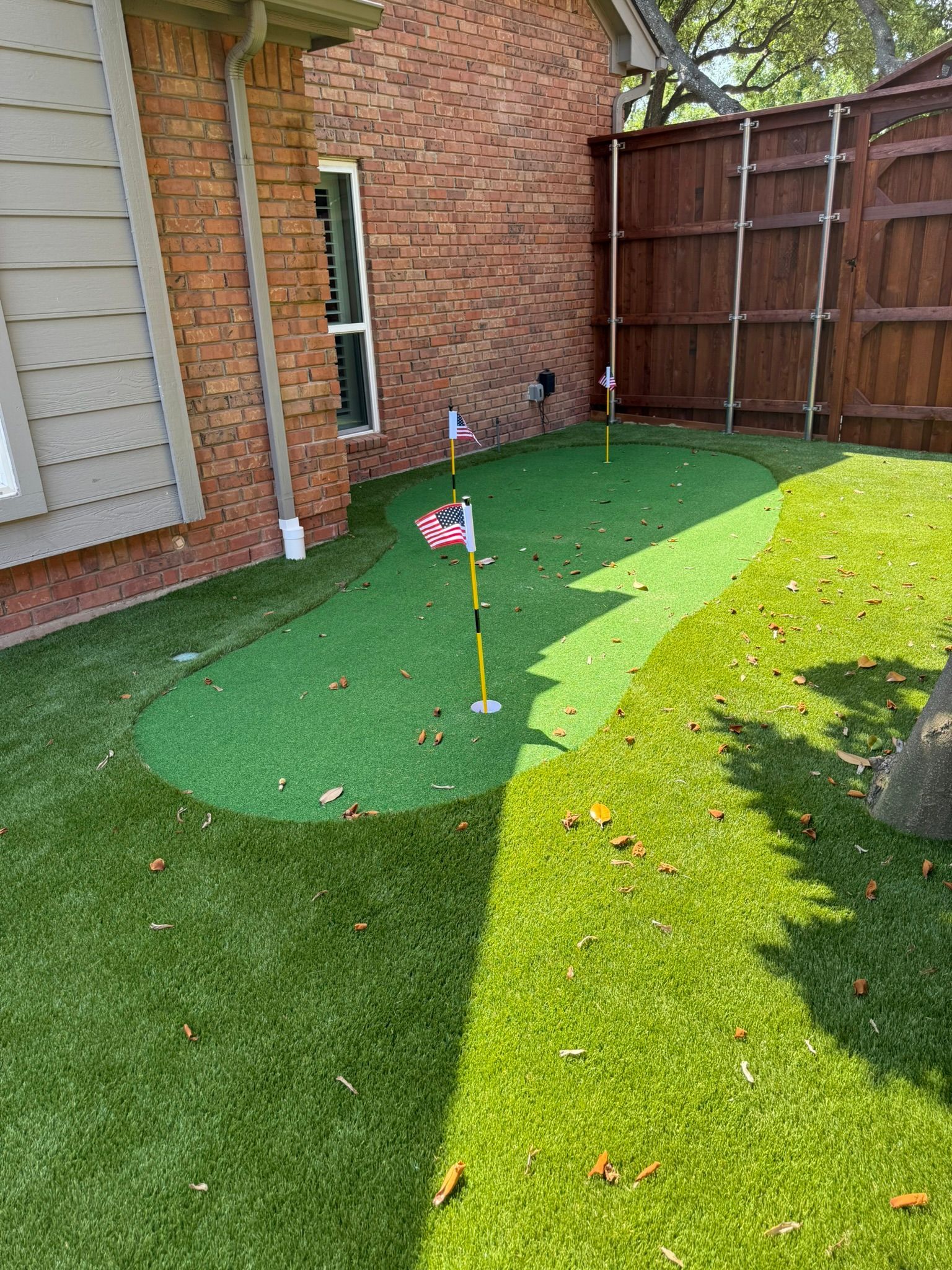 Backyard putting green with flags, surrounded by artificial turf, brick wall and wooden fence.