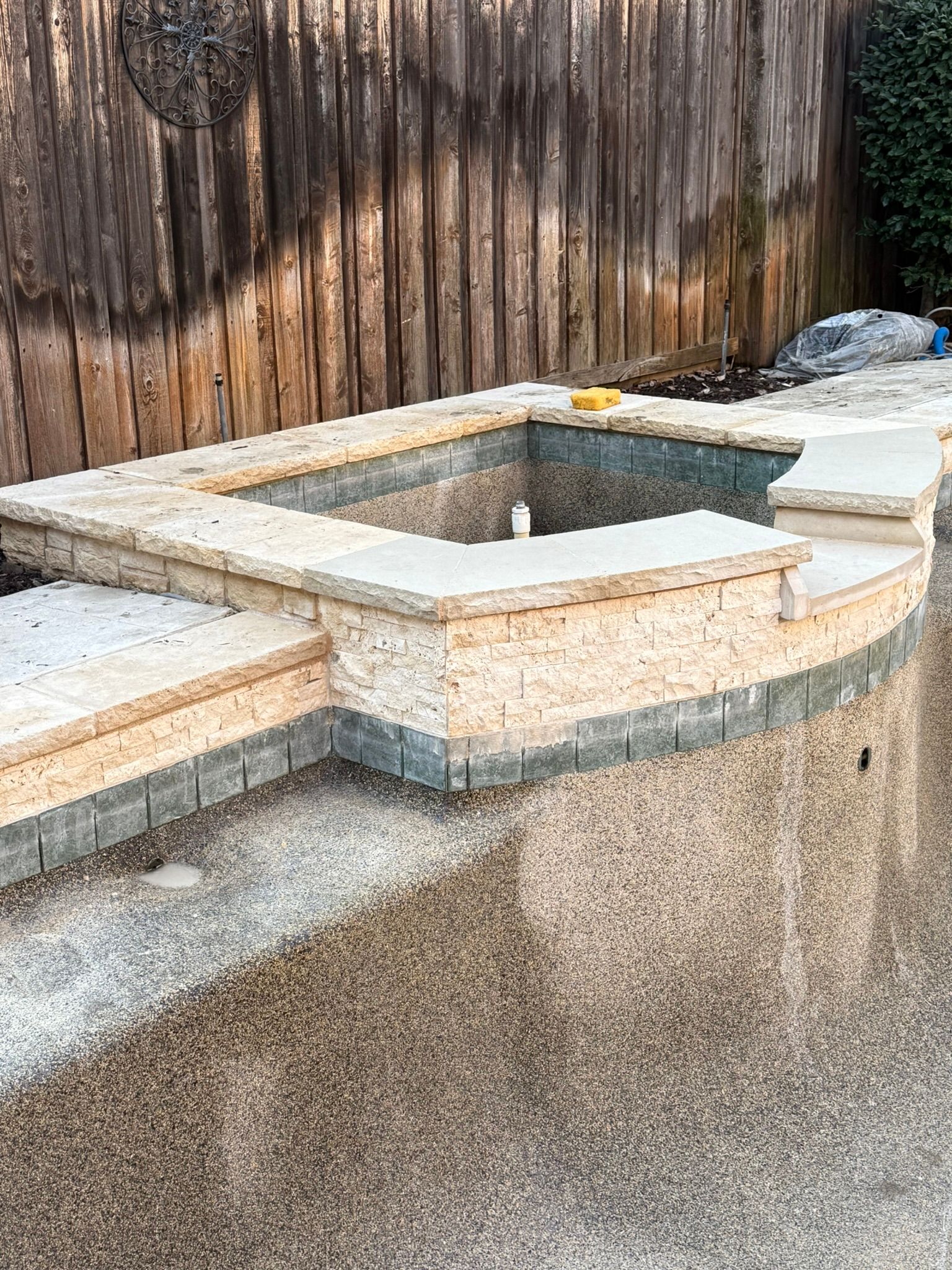 Pool with a built-in spa area under construction; stone and tile edging, wooden fence backdrop.