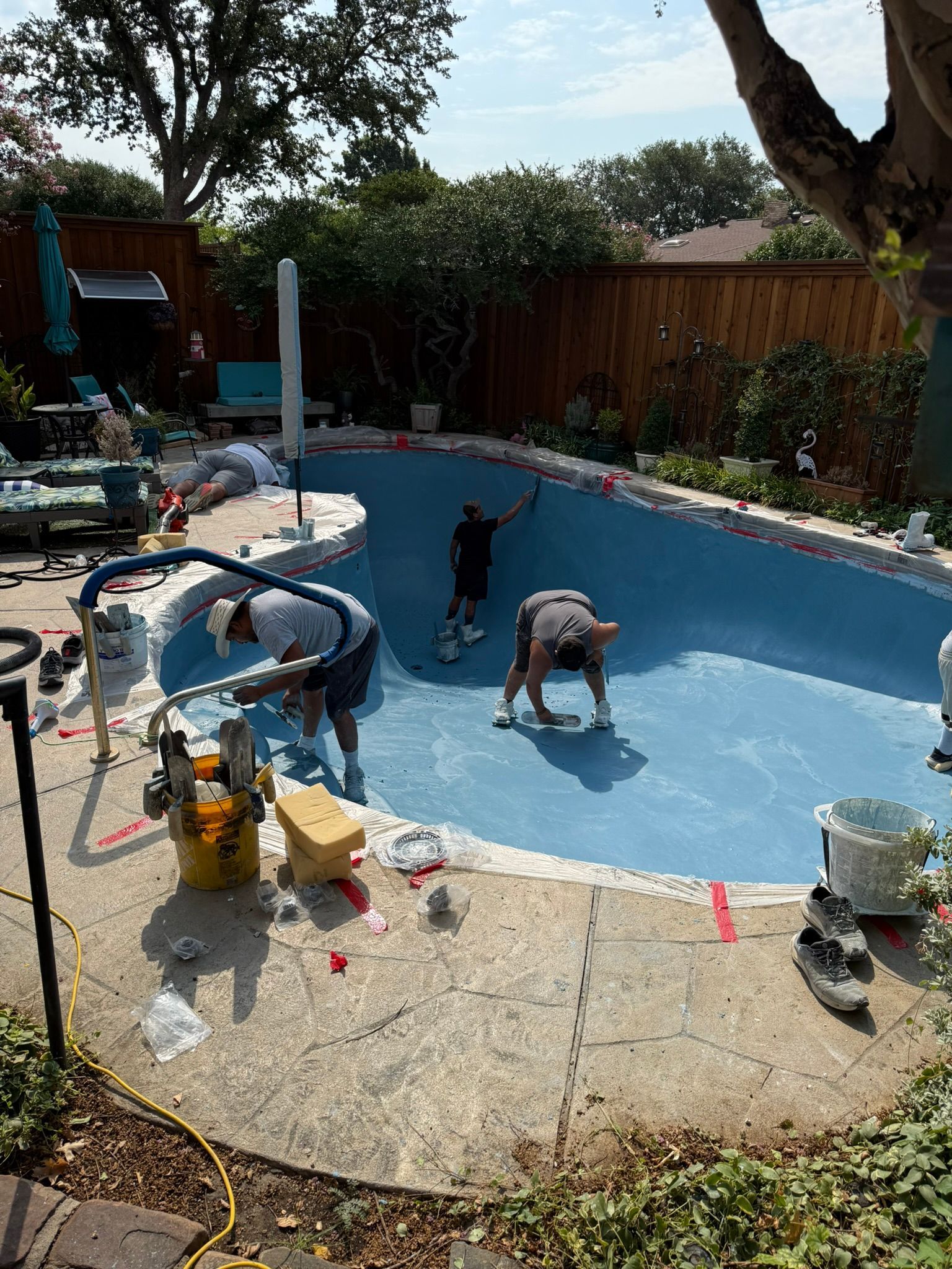 Three people working on the resurfacing of a blue-painted swimming pool in a backyard.