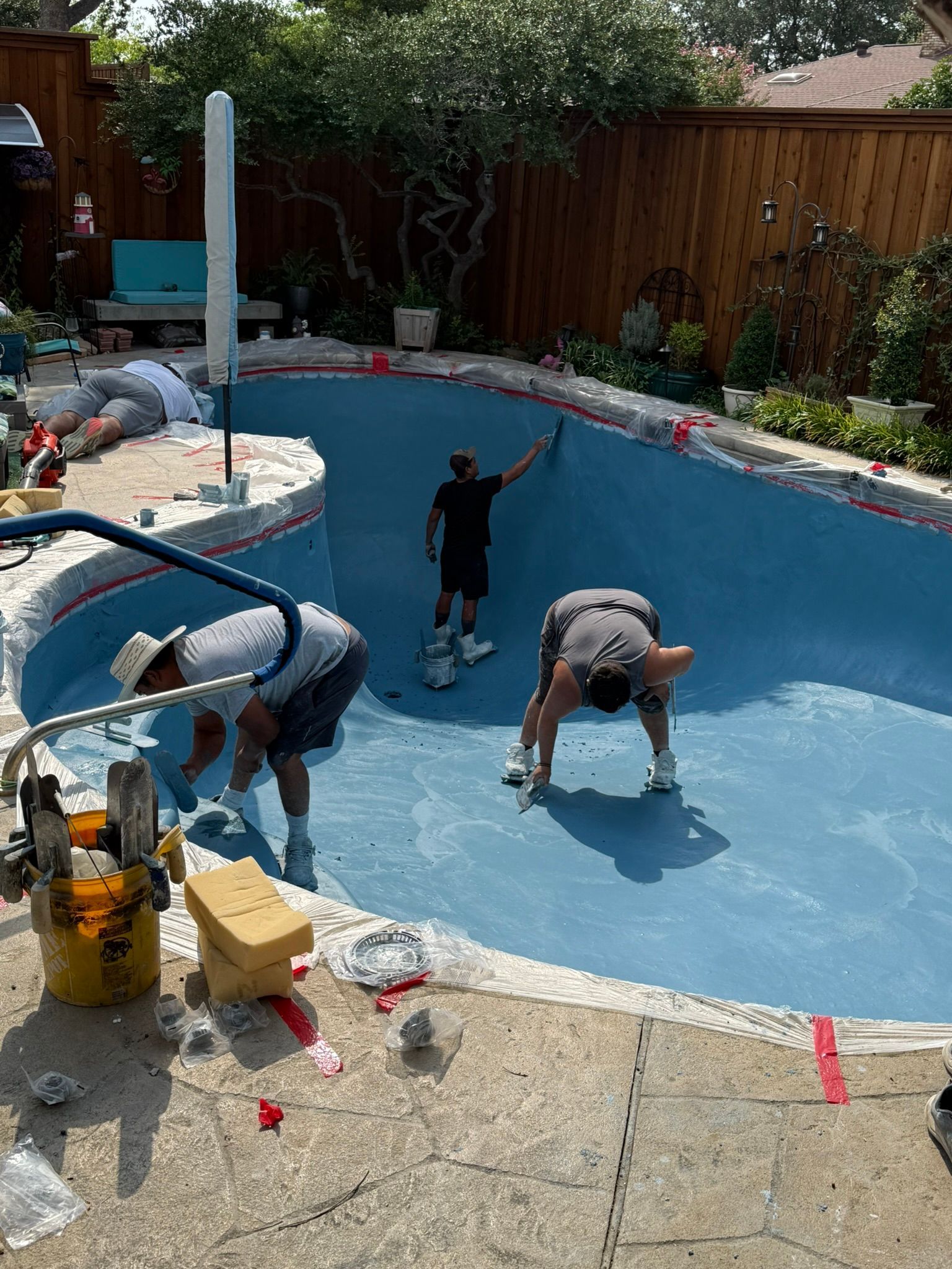 Workers resurfacing a blue swimming pool. They are in the pool, and the pool's edge is being worked on.