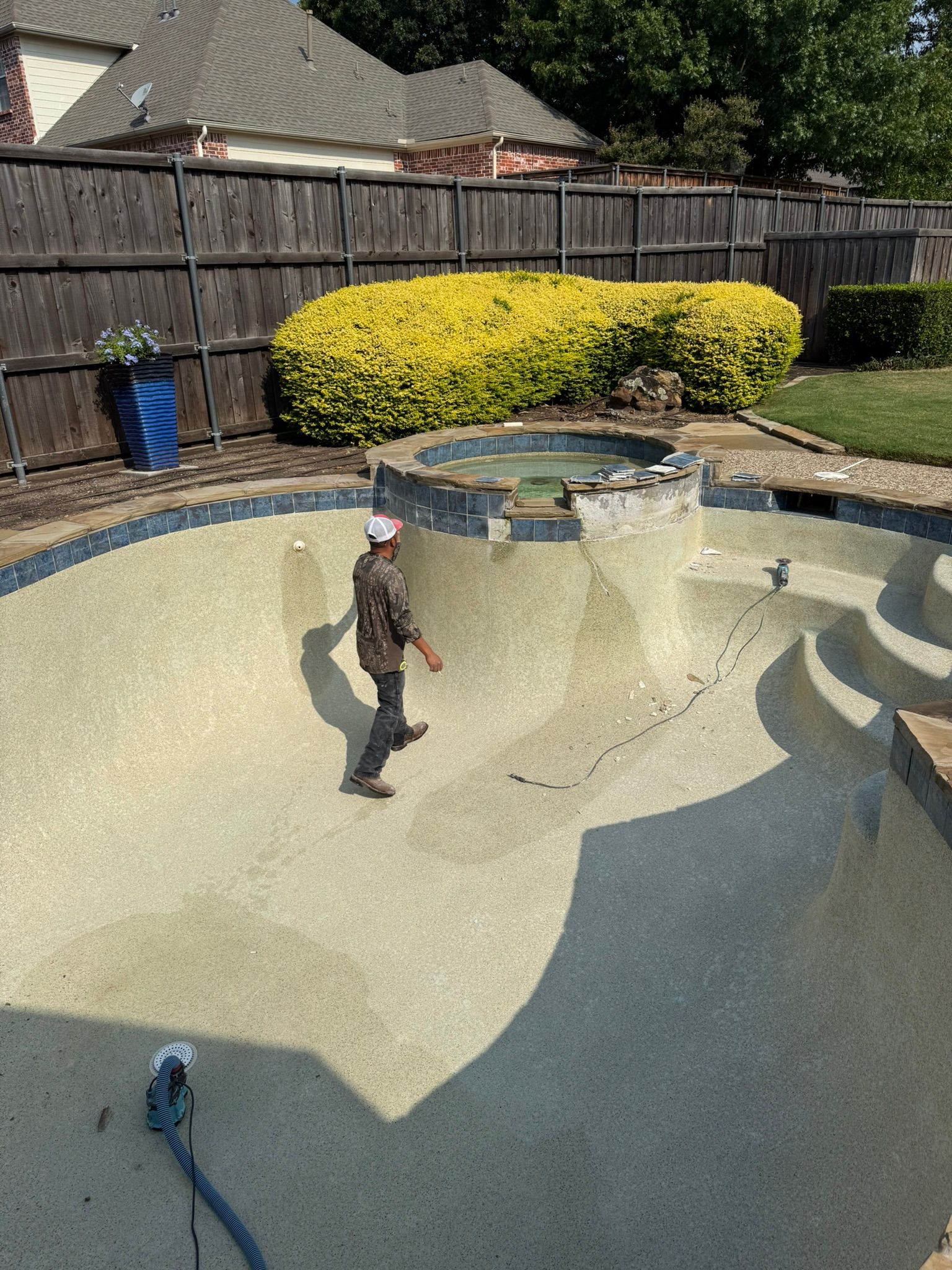 A person walks in an empty swimming pool. The pool is tan with blue tile edges, surrounded by a fence and greenery.