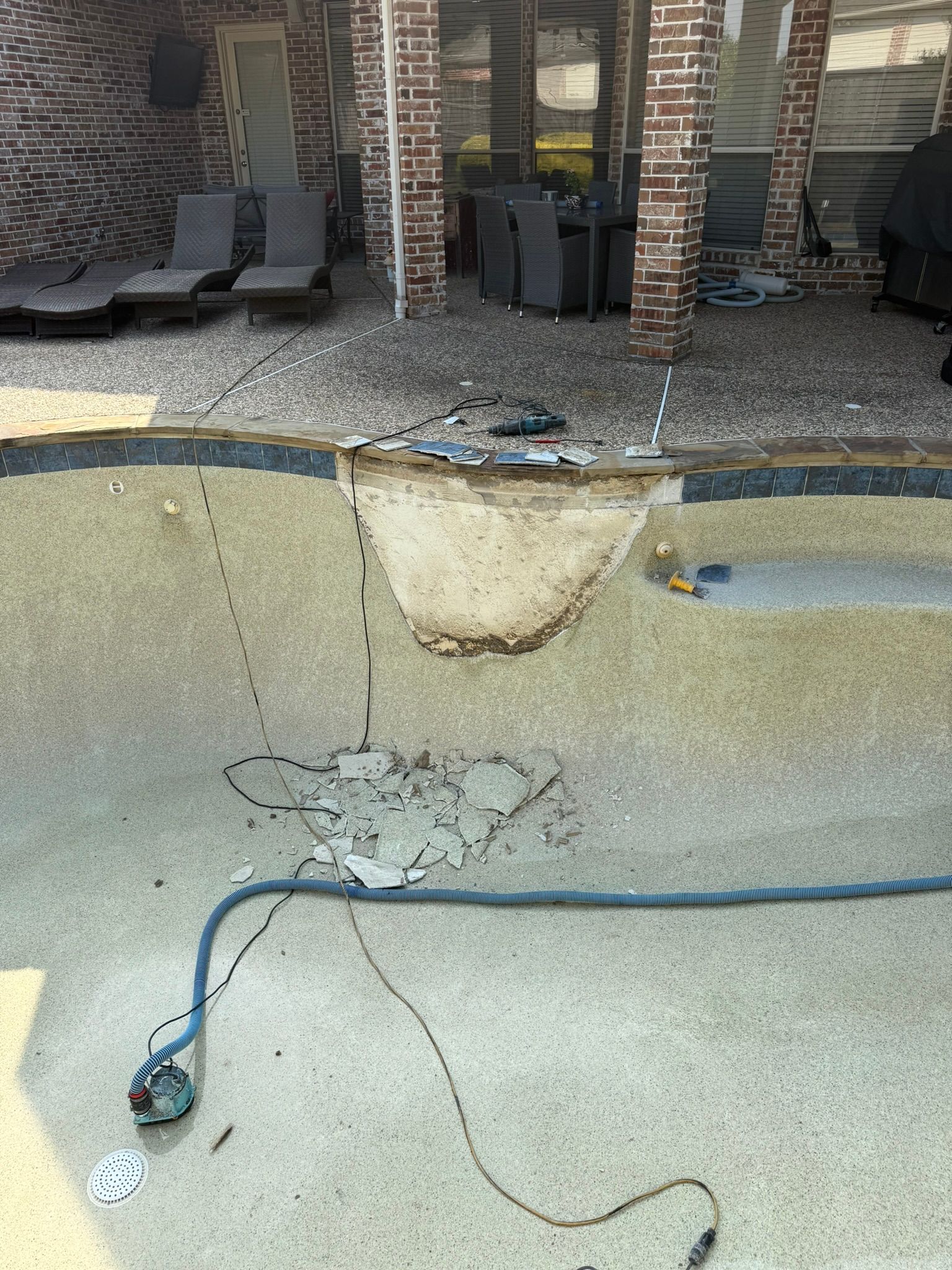 A drained swimming pool with damaged concrete. Electrical wiring and a hose are visible. Patio furniture and a brick structure in the background.