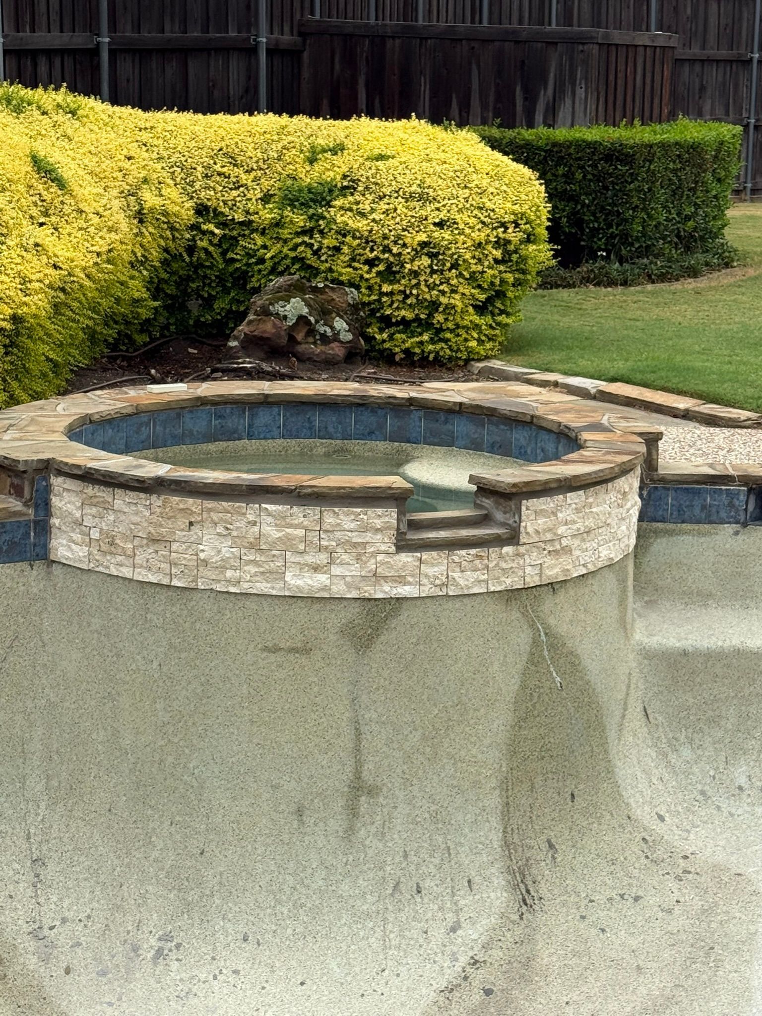 Circular stone hot tub with blue trim, near a lawn and bushes.