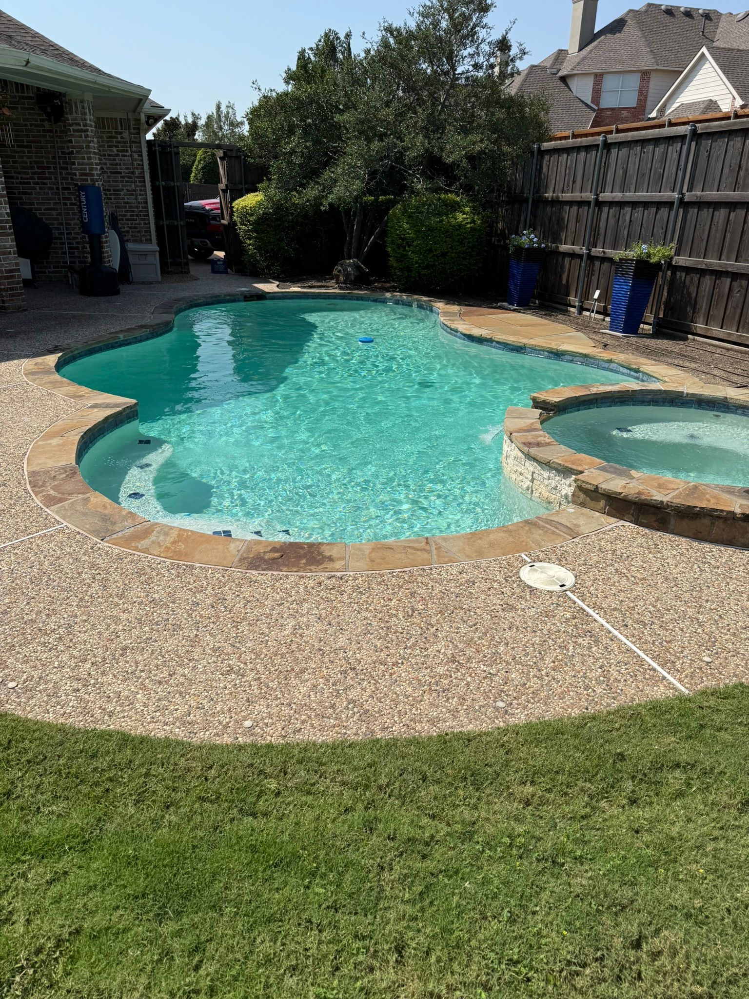 Pool with turquoise water, brown pebbled patio, and hot tub surrounded by green lawn.