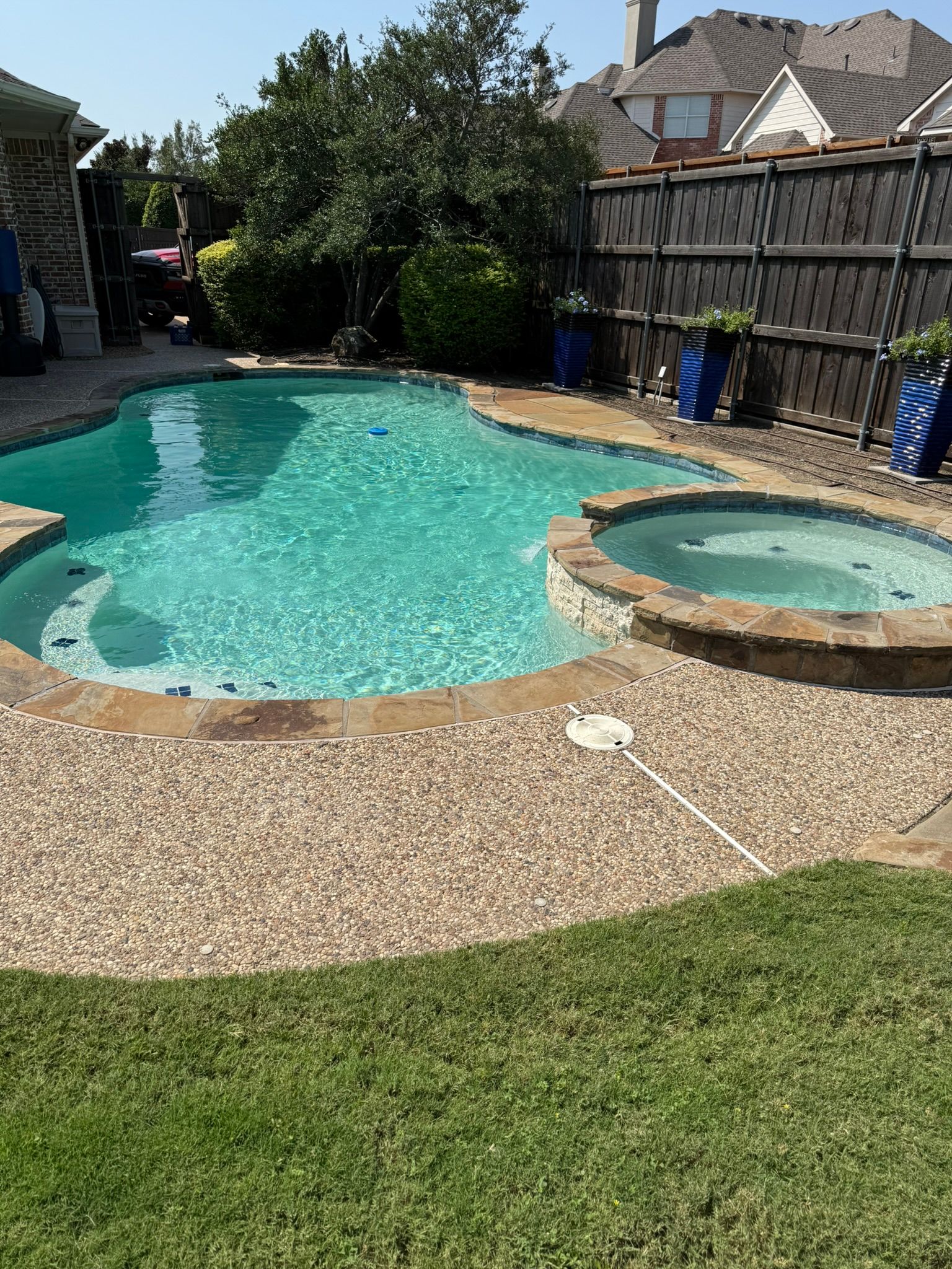 Pool and hot tub with stone surround, blue water, and green grass. Fenced backyard.