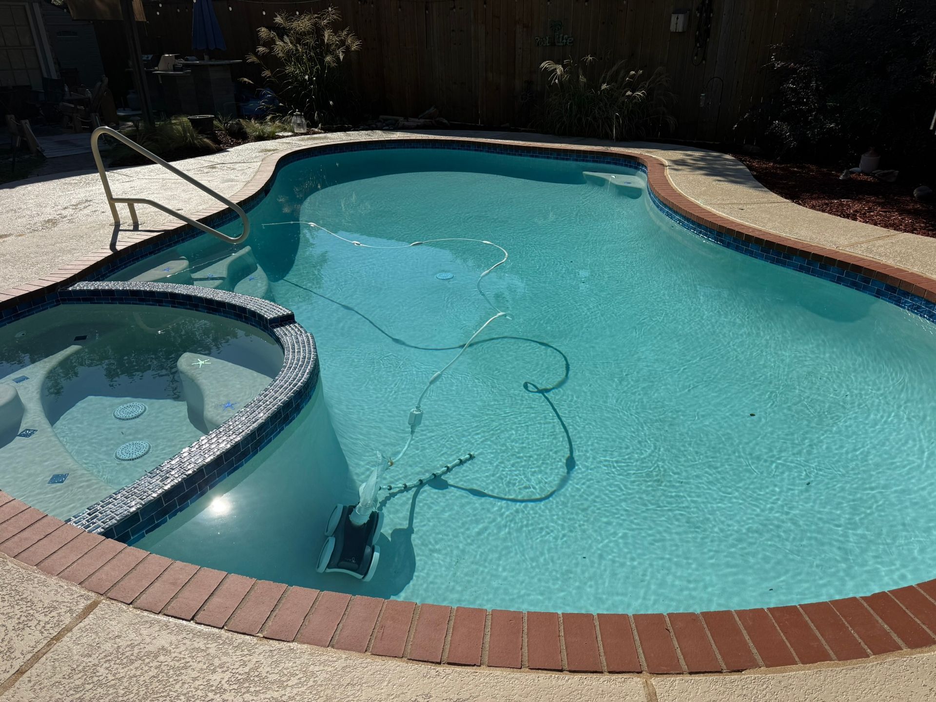 Pool with attached hot tub; blue water, red brick border, concrete deck, and a pool cleaner.
