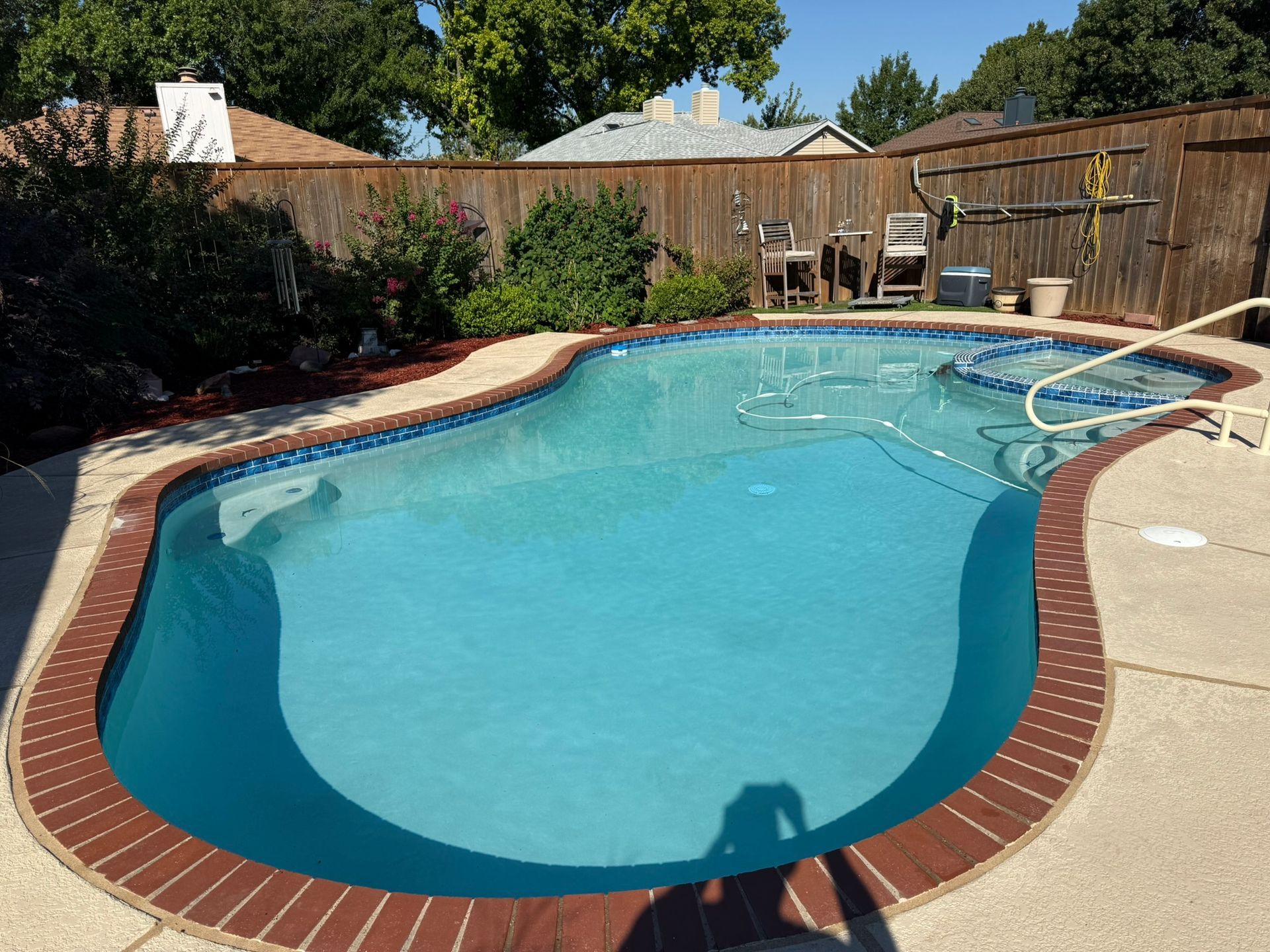 Swimming pool with blue water and brick border in a backyard with wooden fence.