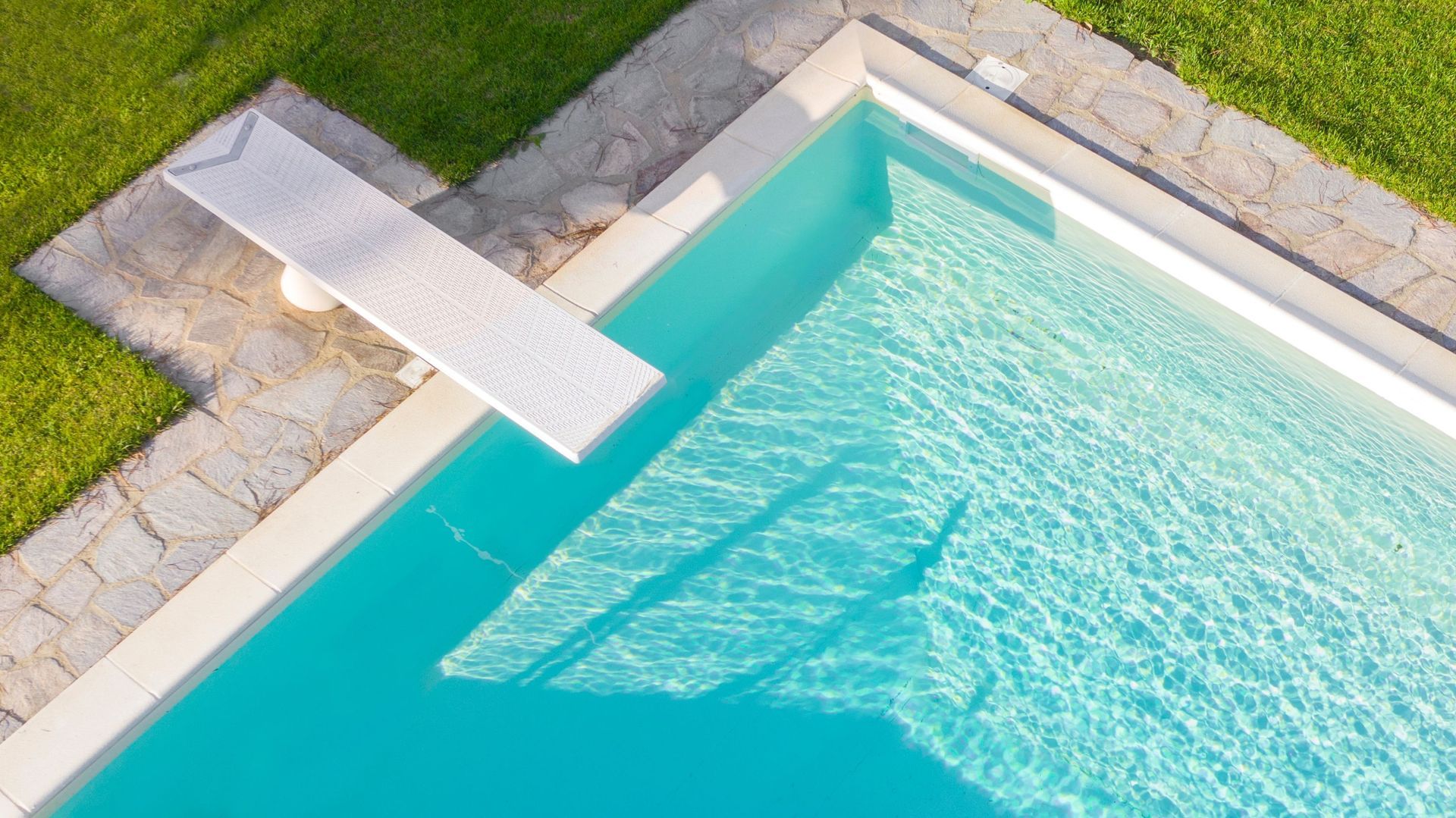 Bright blue pool with a white diving board, stone border, and green grass.