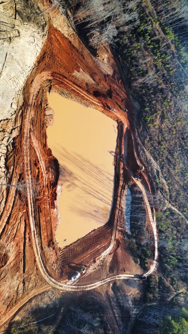 Aerial view of a brown earthen construction site with a muddy water-filled pit, surrounded by dirt roads and trees.