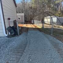 Gravel driveway leading to a shed next to a house, under a chainlink fence and a tree arch.
