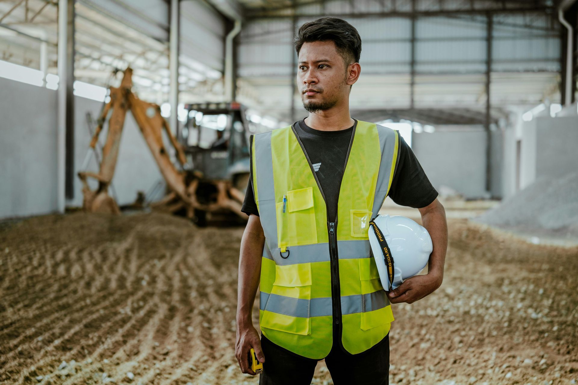 A construction worker wearing a yellow vest and holding a hard hat is standing in a warehouse.