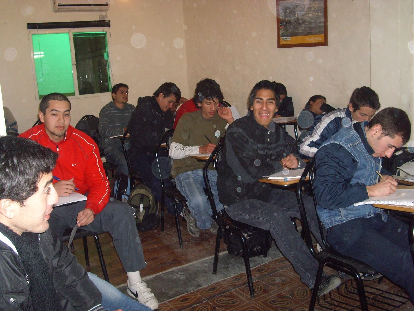 Aula con alumnos escribiendo en sus pupitres. Uno sonríe a la cámara, los demás están concentrados en su trabajo.