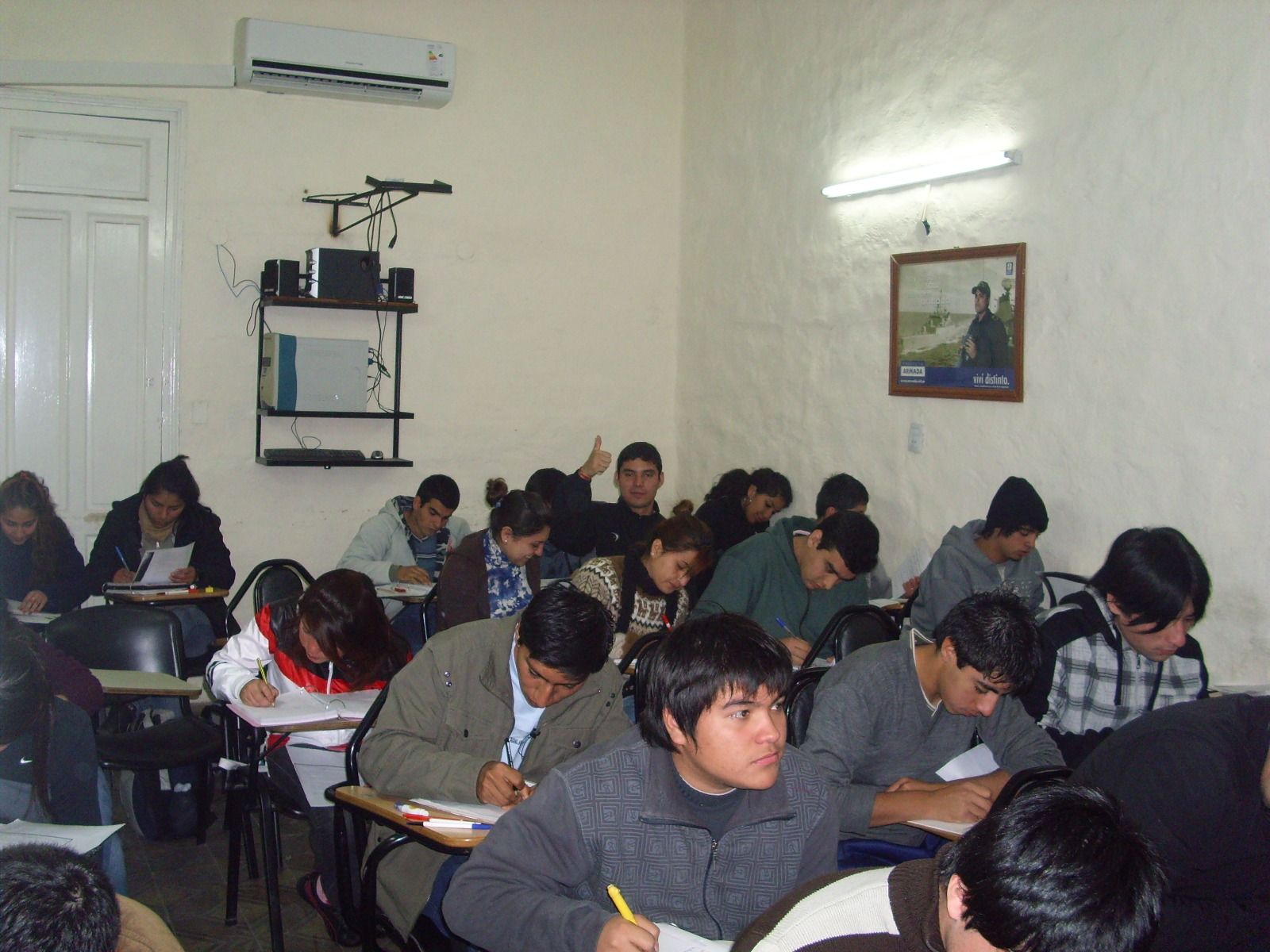 Aula con estudiantes escribiendo en sus pupitres; un estudiante levanta la mano.