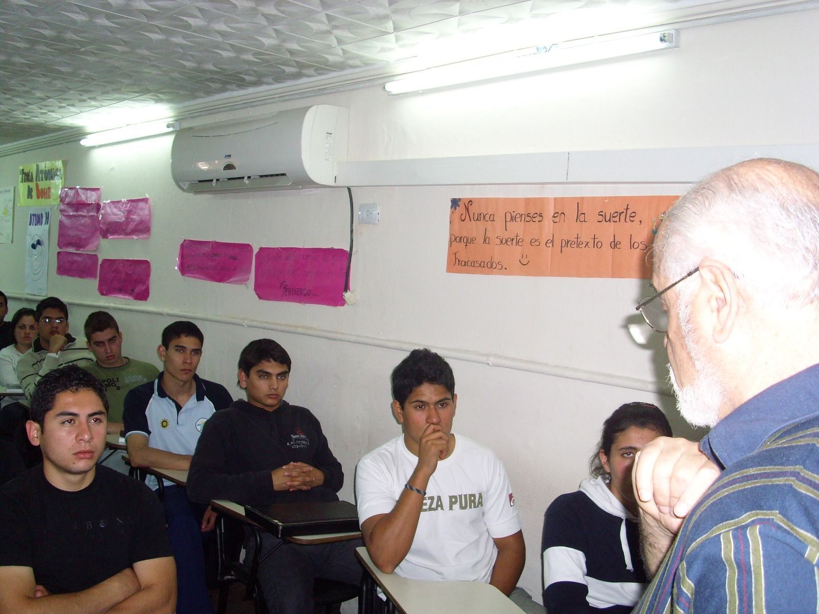 Aula con estudiantes escuchando a un profesor. Los estudiantes están sentados en pupitres; se ve una pizarra blanca.