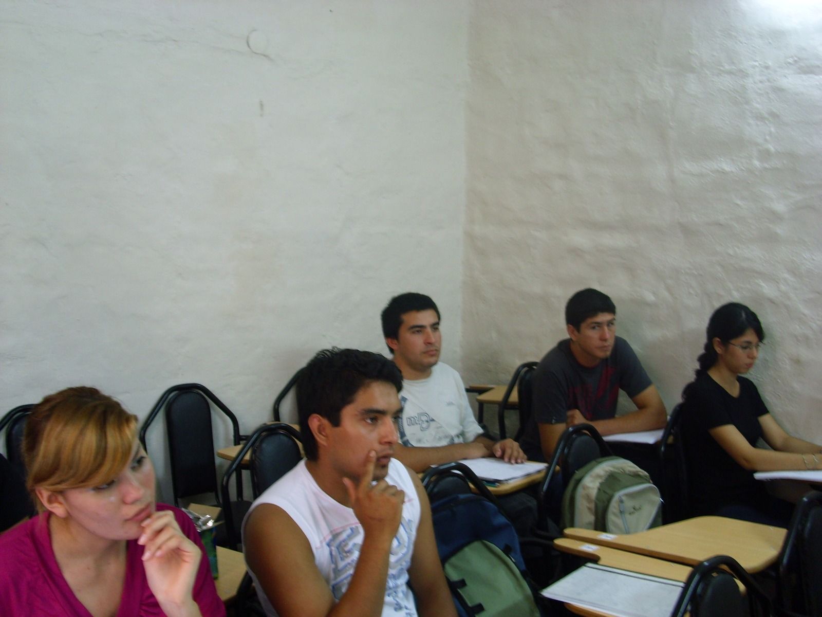 Personas sentadas en pupitres en un aula, mirando al frente. Fondo de pared blanca.