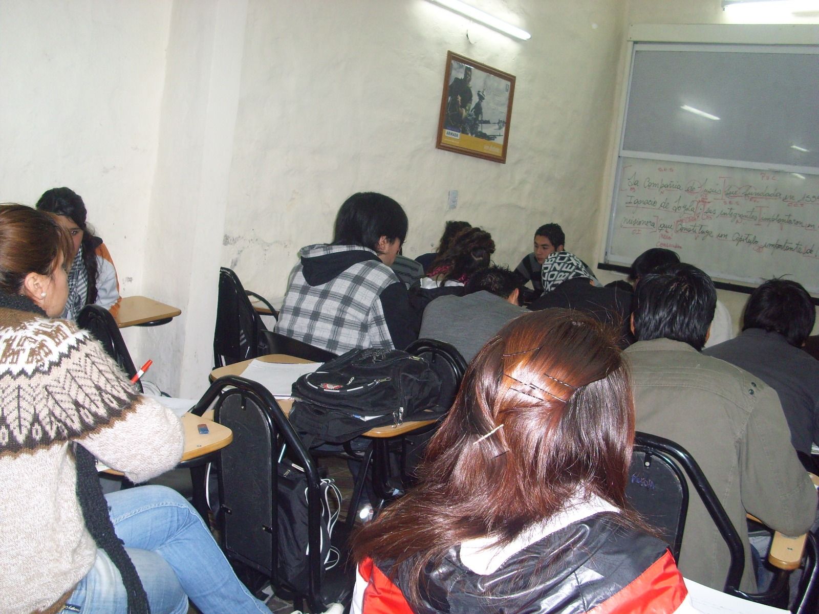 Aula con estudiantes sentados en sus pupitres, mirando hacia adelante. Algunos toman apuntes, otros escuchan o miran al frente.
