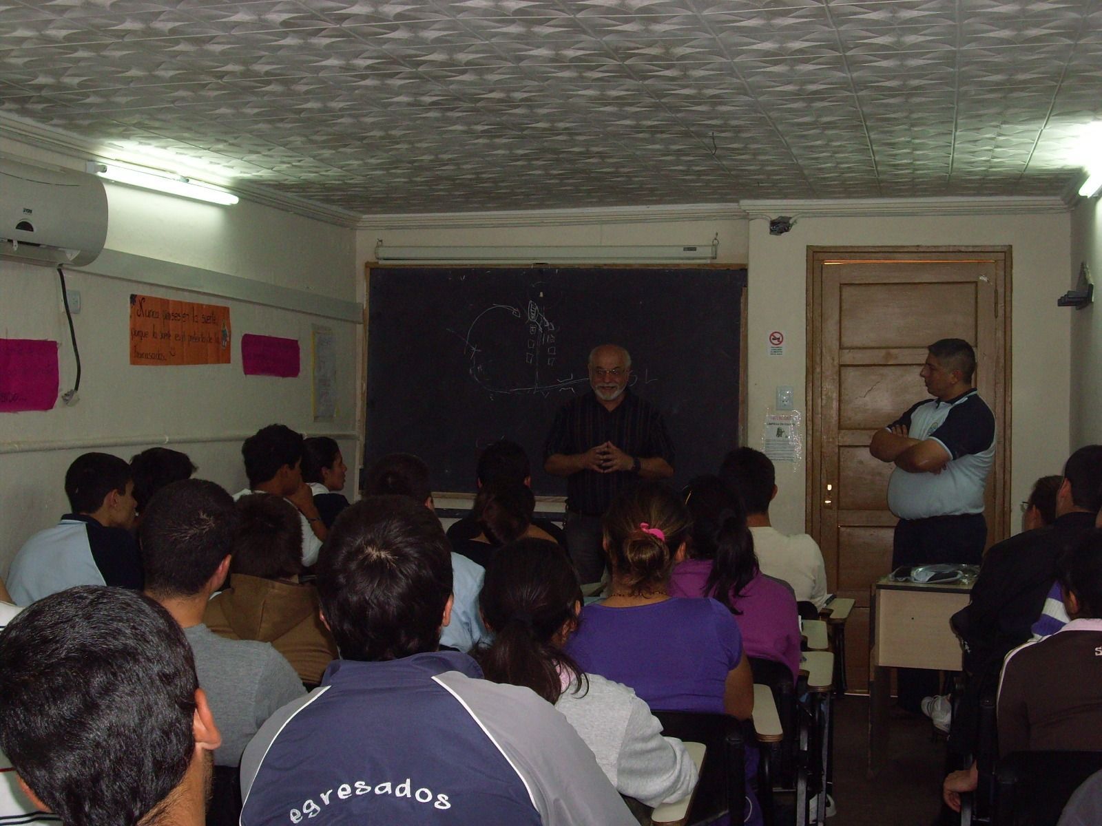 Un hombre está dando una clase a un grupo de estudiantes en un aula, otro hombre está de pie a un lado, dibujando en una pizarra.