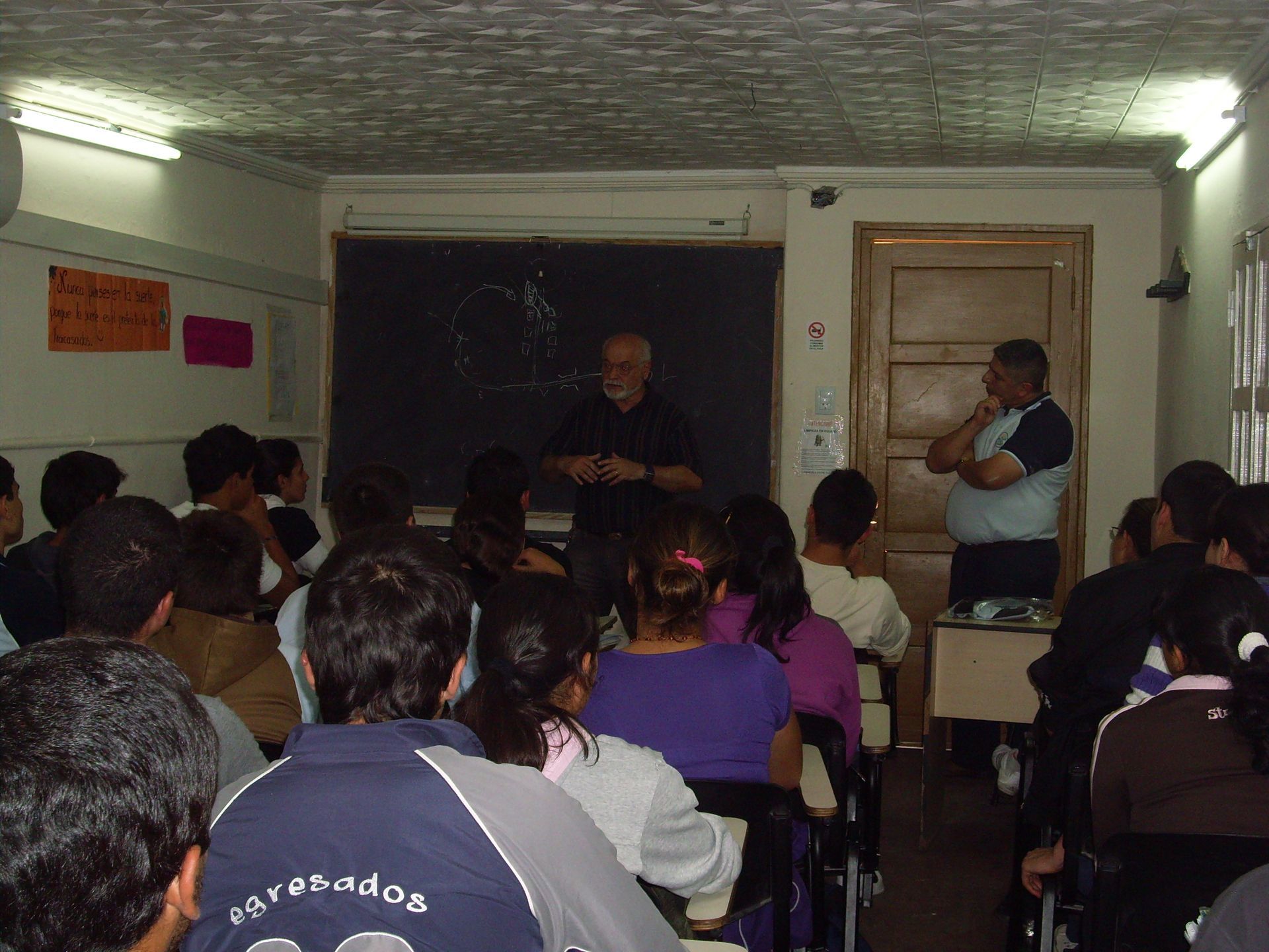 Un hombre da una clase en un aula a un grupo de personas sentadas. Otro hombre permanece de pie cerca de una puerta cerrada, observando.