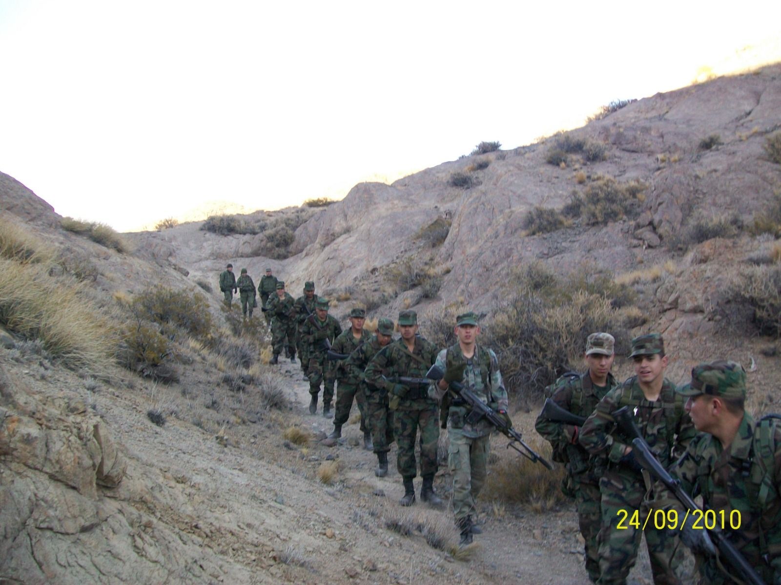 Soldados con uniformes de camuflaje caminan en fila india por un cañón del desierto, fusiles en mano.
