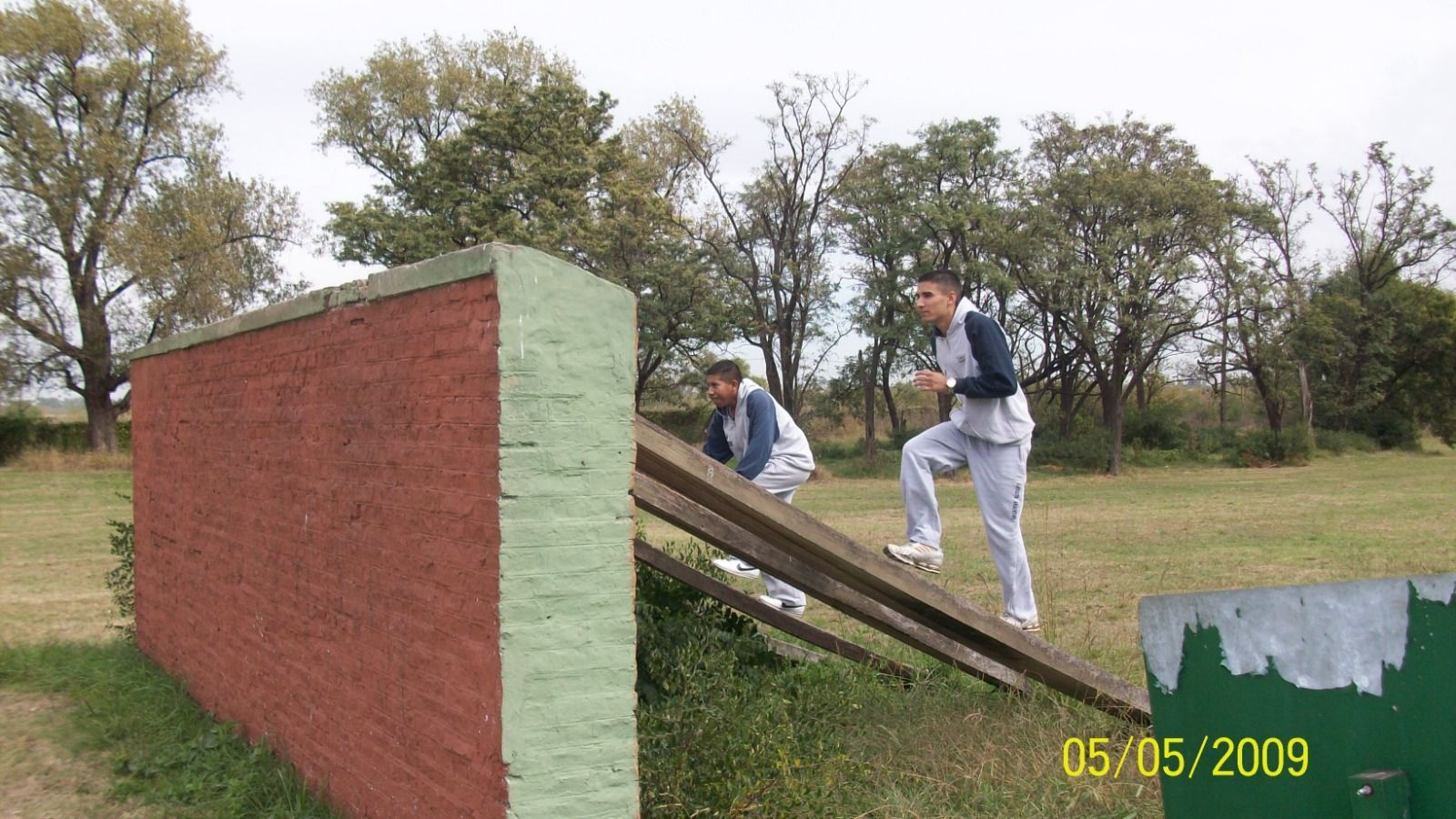 Dos personas escalando un obstáculo de madera en un circuito de entrenamiento al aire libre, con árboles de fondo.