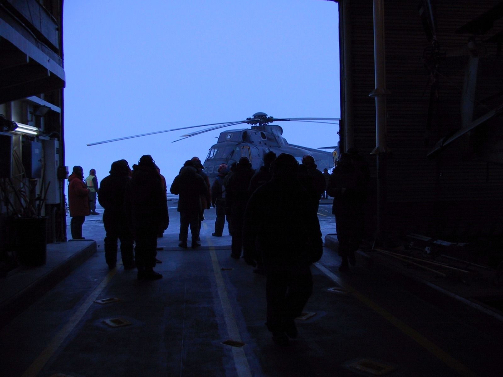 Siluetas de personas caminando hacia un helicóptero sobre una superficie cubierta de nieve bajo un cielo crepuscular.
