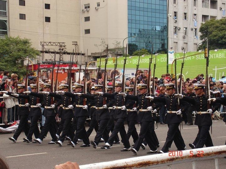 Soldados con uniformes azules marchando en formación, portando fusiles. Edificios y multitud al fondo.