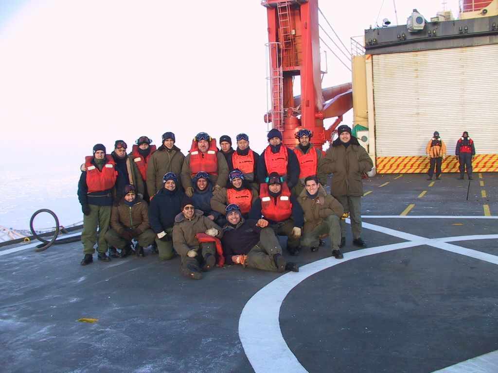 Grupo de personas en la cubierta de un barco con hielo al fondo, vestidas con ropa de invierno y chalecos salvavidas.