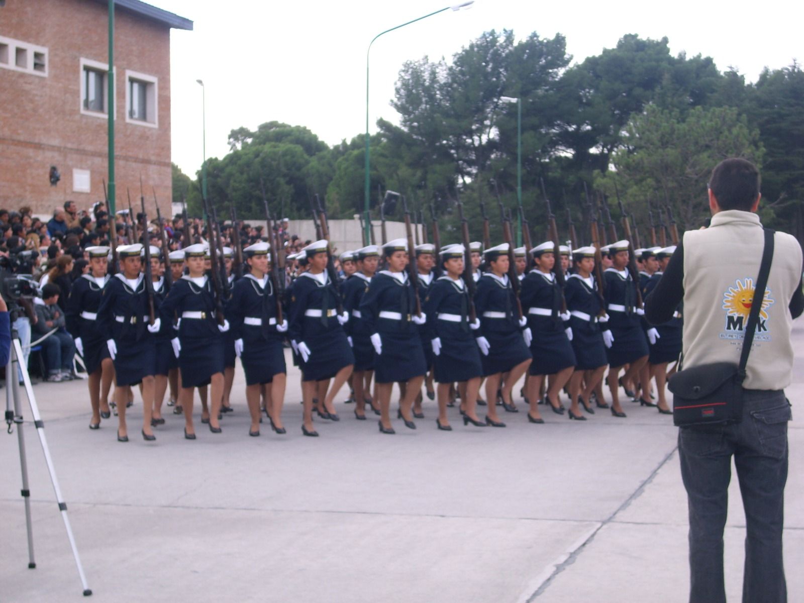 Un grupo de personas uniformadas marchan en formación, portando rifles. Llevan uniformes y gorras de la marina al aire libre.