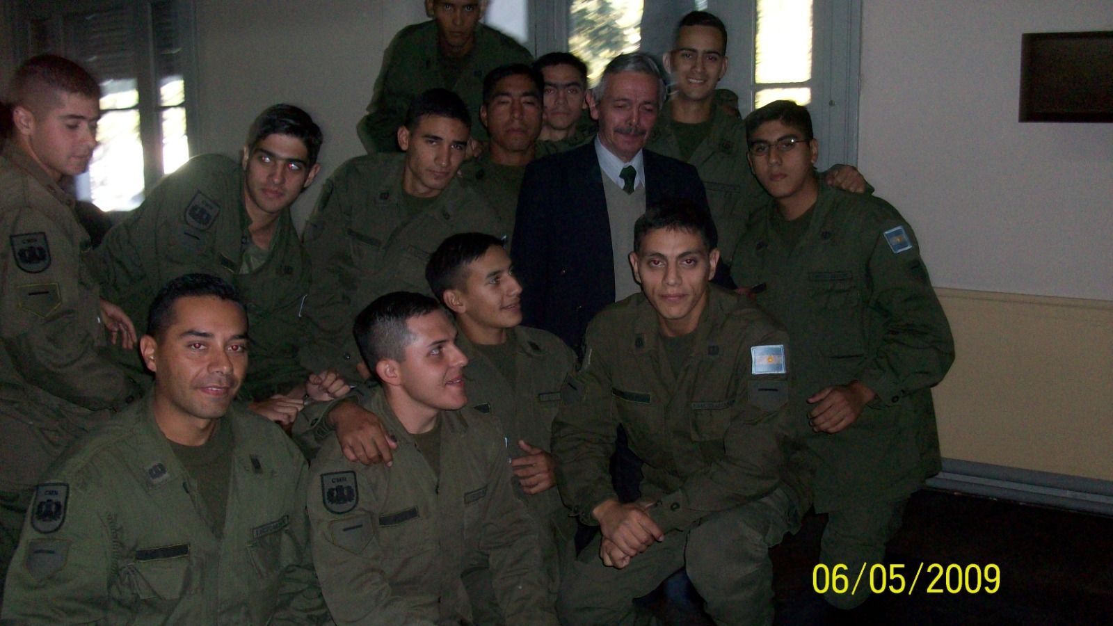 Un grupo de hombres con uniformes militares posan con un hombre de traje, en el interior, cerca de una ventana.