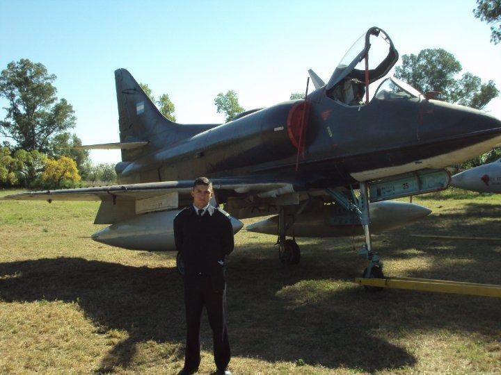 Un hombre uniformado está de pie frente a un avión A-4 Skyhawk de color gris oscuro en un campo de hierba.