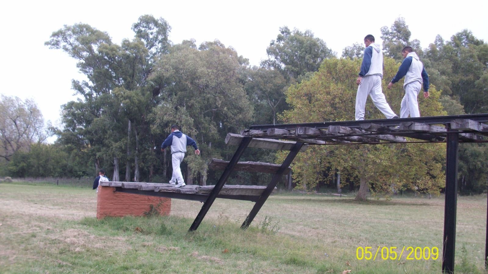 Tres personas con uniformes blancos caminan sobre una pista de obstáculos de madera al aire libre, junto a una estructura de ladrillo y árboles.
