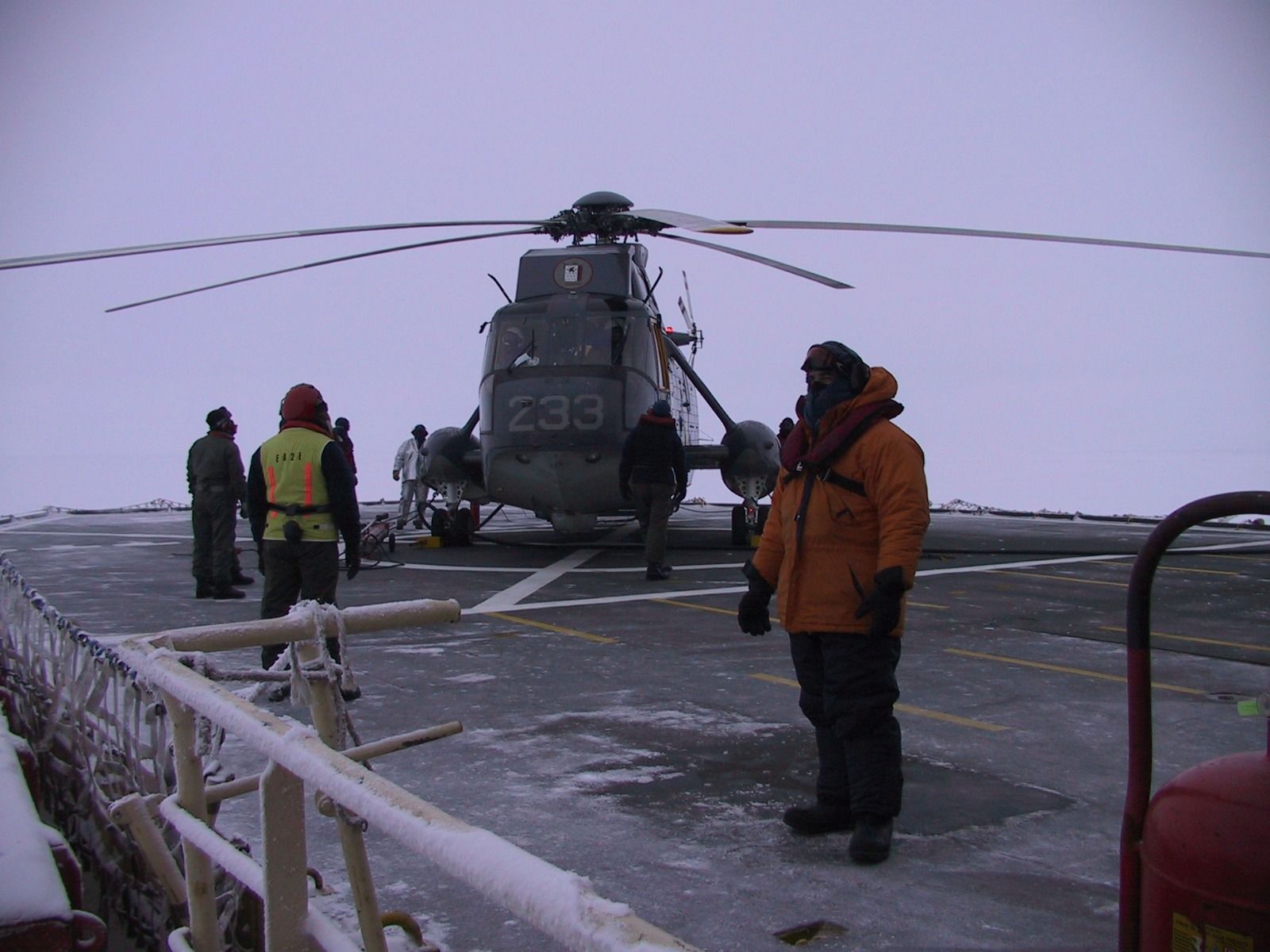Helicóptero sobre cubierta nevada con miembros de la tripulación. Un hombre con abrigo naranja está de pie en primer plano.