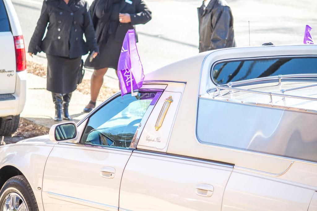 Beige hearse with people in black near it.
