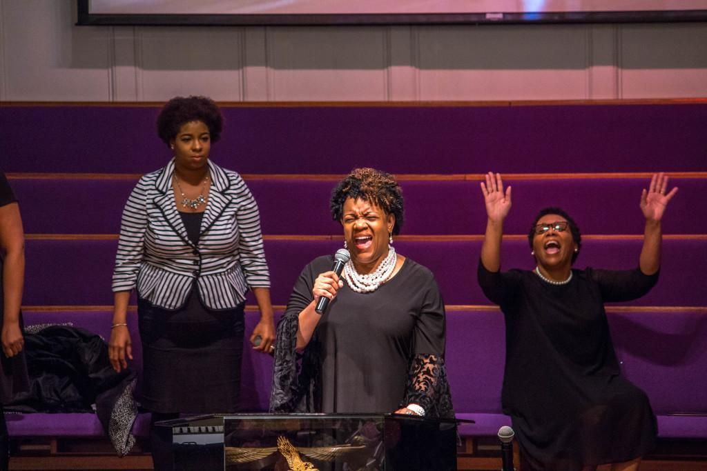 Choir singing on stage at a church. Woman sings into mic, others raise hands and look joyful. Purple backdrop.