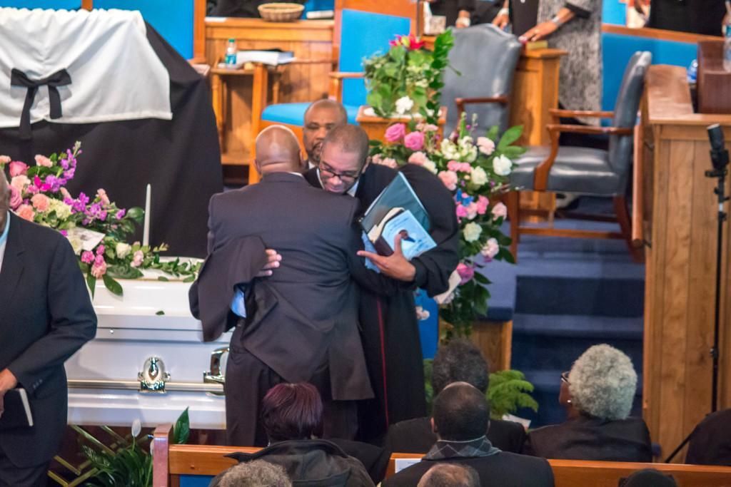 Mourners hug at a funeral. A white casket, flowers, and a preacher with a photo stand in a church.