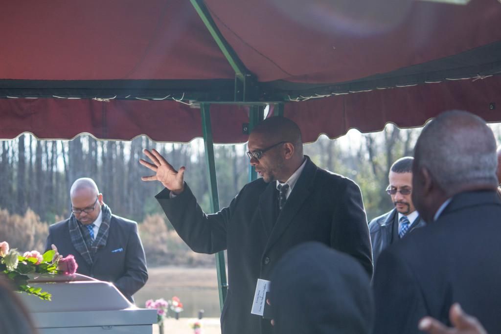 Man speaking at a graveside service, gesturing with his hand. Others stand nearby.